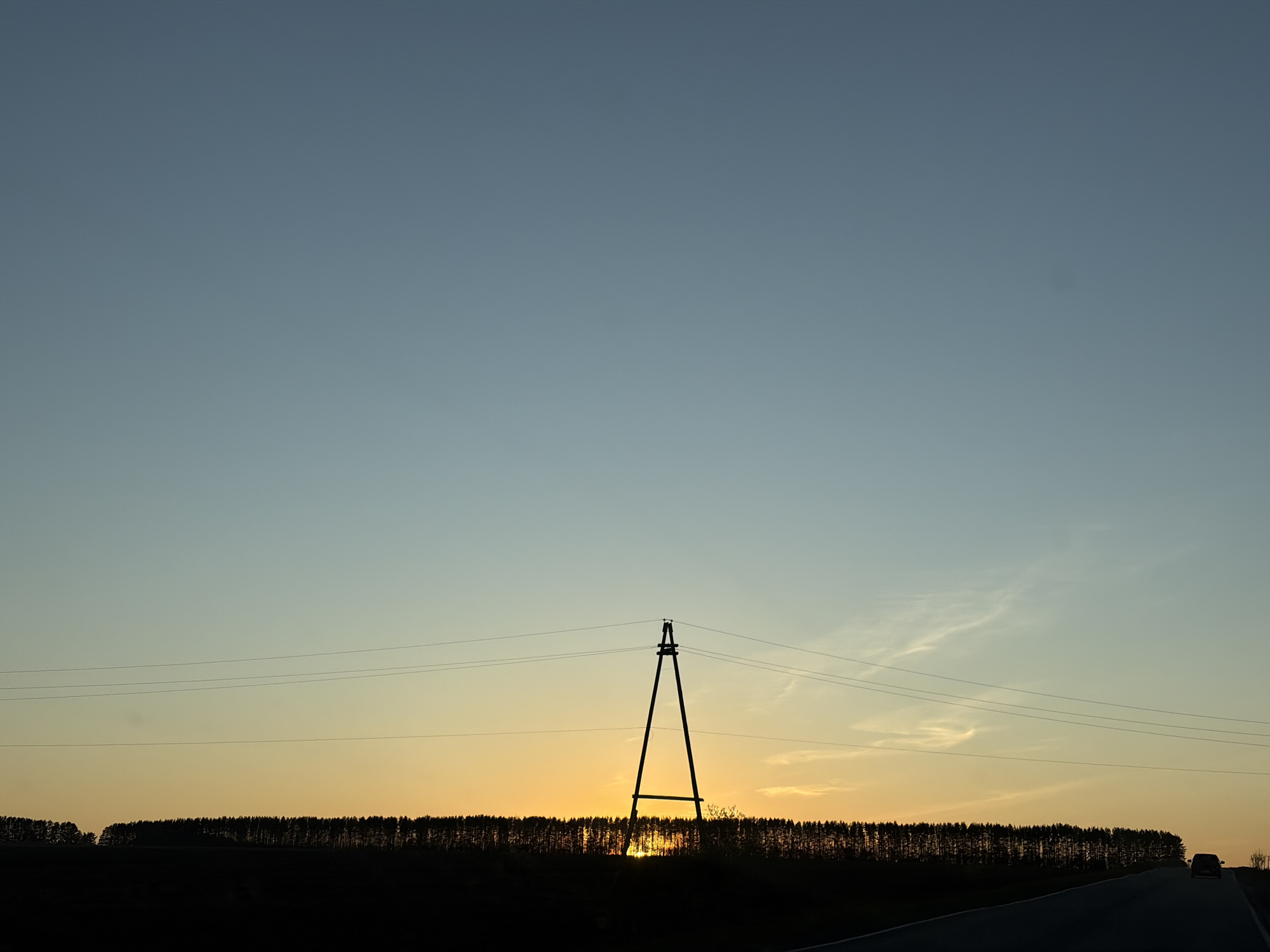 A landscape scene shows a silhouette of a power transmission tower and a dense line of trees silhouetted against a sunset. Wires are strung from the tower. The sky transitions from a light blue at the top to a warmer orange near the horizon. A road runs along the bottom right of the image. There is one car visible on the road, and the location is indicated to be Nizhnii Vishur, Russia.