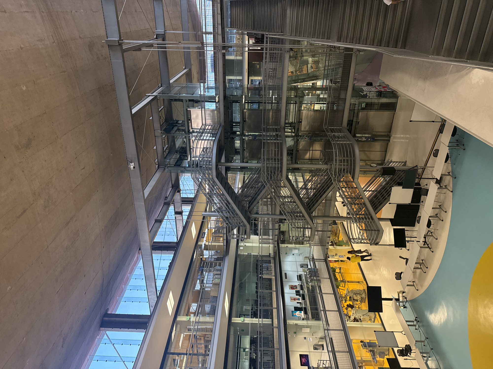 The image depicts the interior of a modern, multi-story building in Mexico City, Mexico. The architectural style features exposed steel beams, glass walls, and staircases. The interior includes a light-blue floor with a yellow circle. The space is organized with tables, chairs, and computer monitors. In the center of the building are various metal stairs, platforms, and caged elevators. People can be seen walking around the space, with one individual in a yellow shirt. There are various objects, including machinery and research equipment, behind glass walls. Overall, the environment is an open, modern, and functional space.