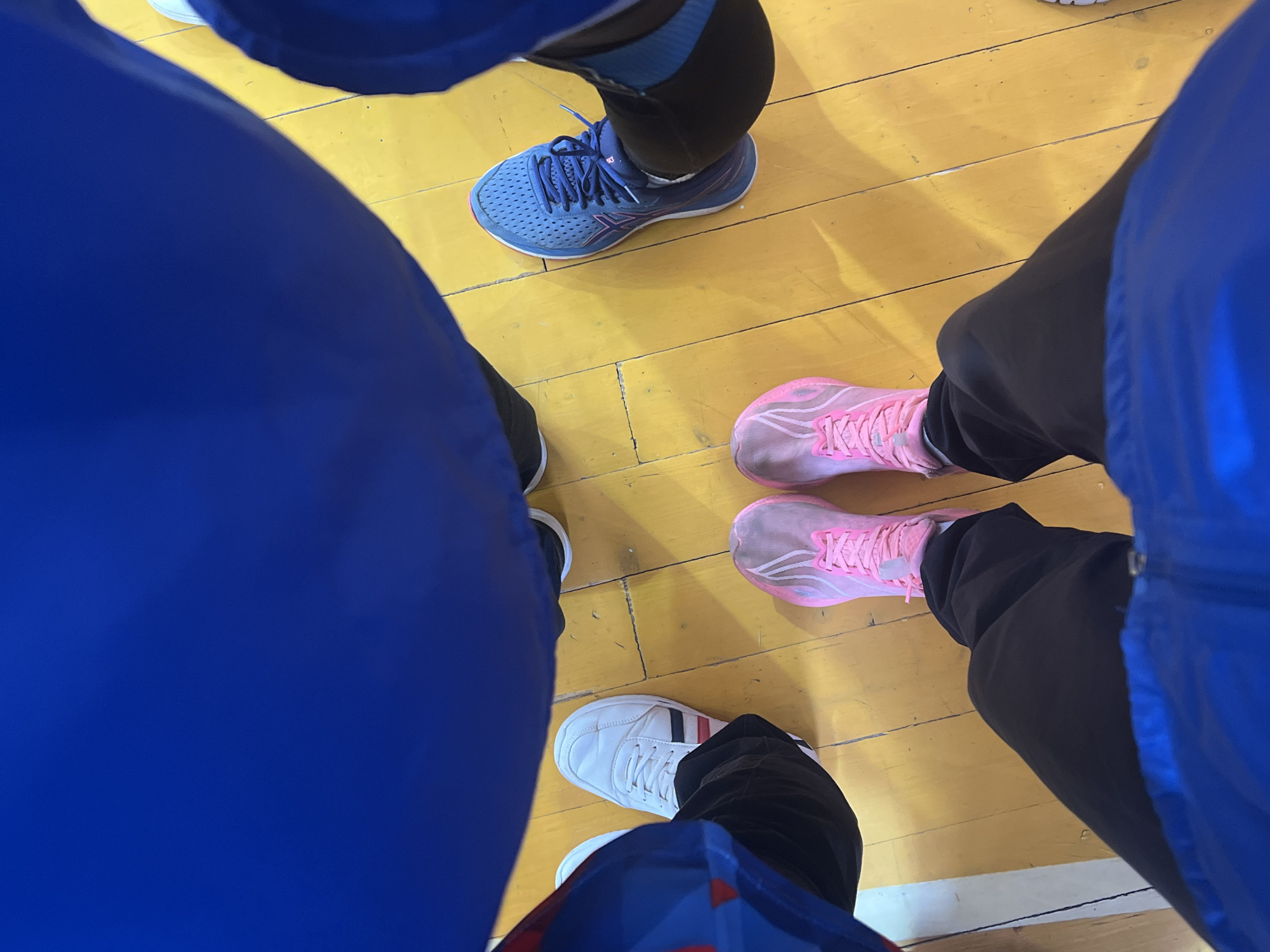 A high-angle shot depicts the feet of four individuals standing on a wooden floor, potentially a gymnasium, located in Chaikovsky, Russia. One individual wears blue sneakers with dark socks or leggings. Another individual has pink running shoes and dark pants. A third person wears white sneakers with red and blue stripes. The clothing of the individuals is predominantly blue and black. The floor is light yellow with distinct planks. No specific actions or interactions are clearly depicted beyond their proximity.