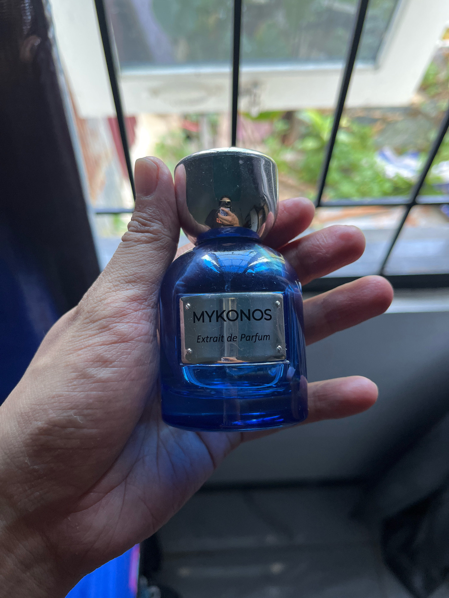 A hand holds a blue glass perfume bottle with a silver cap and a silver label that reads "MYKONOS Extrait de Parfum." The background appears to be an outdoor setting with foliage visible through a window with black bars. The image quality is moderate, with some blur and lighting challenges. The location is Tungkaran Pangeran, Indonesia.