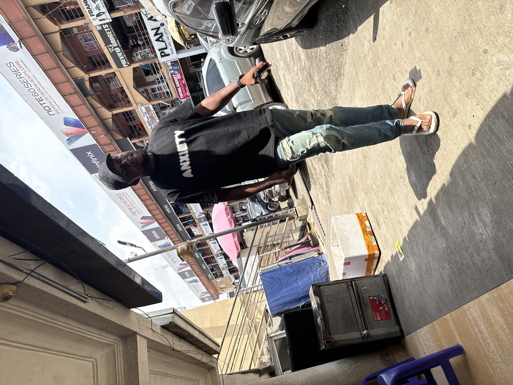 An outdoor, sunlit scene captures a man standing on a concrete sidewalk in what appears to be a bustling commercial area, likely in Abuja, Nigeria. The man wears a black t-shirt with the word "ANXIETY" printed in white, distressed blue jeans, white sandals, and a patterned cap. He holds a dark smartphone in his right hand.
The setting features multi-story buildings, potentially housing electronics and gadget shops, under a bright, clear sky. Strong shadows indicate a sunny day, likely mid-day or afternoon. Behind the man, a silver car is parked on the street. Numerous commercial banners are prominently displayed on the buildings. One large banner advertises "Infinix NOTE60 SERIES READY STEADY | EXPLORE," detailing features like "Snapdragon 7s Gen 45G | Rear Matrix Display / 90W 6500mAh." Other visible signs include "PREVIEW GADGETS," "TOPVAL GADGET PLAN PHONES APPLE AND LAPTOPS," and a partially visible "FRANK." On the ground, various objects like shipping boxes, a blue tarp, and a purple plastic stool are scattered near the buildings. Metal railings and a surveillance camera are also visible elements of the urban landscape.