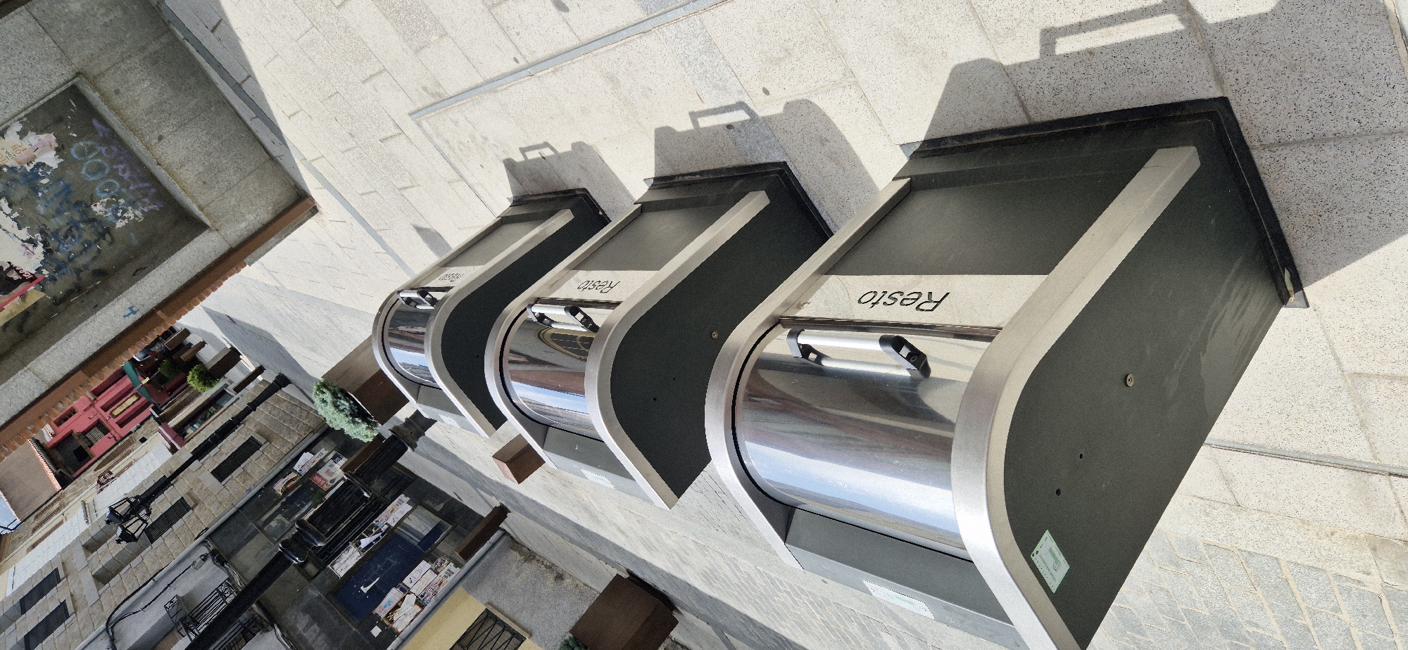 In an outdoor urban setting, four identical, modern trash receptacles are lined up diagonally on a light-colored paved walkway. Each receptacle features a dark gray base with a curved, silver-toned metallic top. A silver handle is centrally positioned on the curved top of each unit, suggesting an opening mechanism for waste disposal. The word "Resto" is inscribed in black, vertical lettering on the side of each receptacle. The scene is illuminated by sunlight, casting distinct shadows from the receptacles onto the pavement. In the background, a narrow street is visible with buildings of varying architectural styles and colors, including a red building and structures with visible windows and balconies. There are also streetlights and potted plants present in the background. The ground is paved with rectangular tiles, and a thin metal strip runs vertically along the right side of the frame. There are no people or animals present in the image.