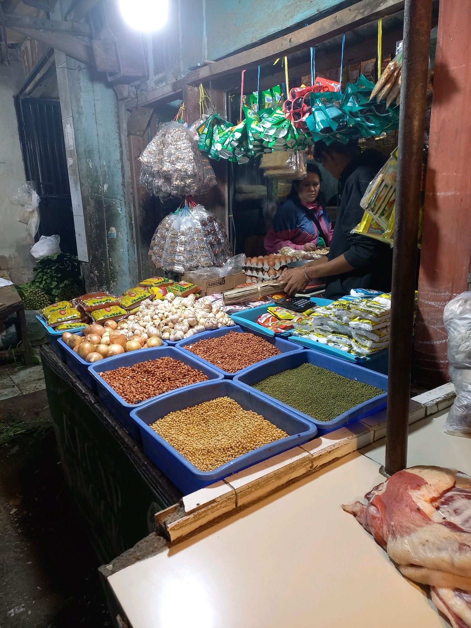 This image depicts a street-side market stall in Bandung City, Indonesia. The stall is well-stocked with various food items. In the foreground, several blue plastic trays hold an assortment of dry goods: peanuts, mung beans, and other legumes. Behind these, onions and garlic are displayed. Further back, shelves are filled with packaged goods, including what appear to be snacks and spices. A woman in a blue and pink jacket is seated behind the counter, and a man in a dark hoodie stands next to her, interacting with the products. Eggs are also visible in trays on a shelf. Hanging from the stall's overhead structure are packages of what look like candies or small food items. To the right, a portion of raw meat is displayed on the counter. The overall environment suggests a bustling local marketplace, likely operating in the evening, given the artificial lighting. The stall appears to be constructed from wood and metal, with a slightly weathered appearance.