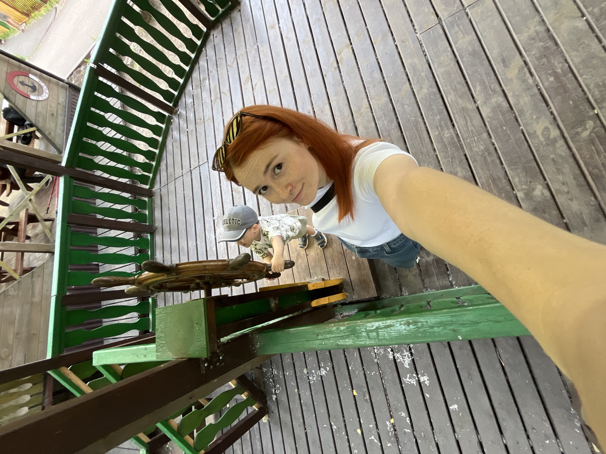 In Chaikovsky, Russia, a woman with red hair and sunglasses on her head is taking a selfie on a wooden platform. She is wearing a white t-shirt and denim shorts. Beside her, a young boy in a cap and a patterned t-shirt is interacting with a ship's steering wheel. The platform has wooden planks and is bordered by a green and brown railing. The scene appears to be part of an outdoor recreational area, possibly a playground or a park.