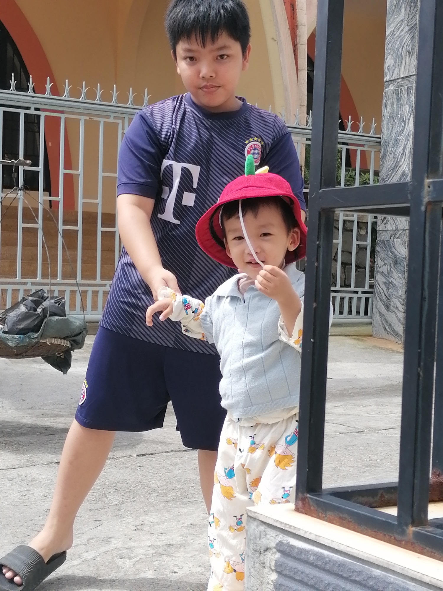 Two young males are pictured outdoors in front of a building with an arched entryway and a decorative fence. The older male, on the left, wears a dark blue shirt with white stripes and a white "T" logo, and dark blue shorts. He is holding a black plastic bag and a woven basket on his left side. The younger male, on the right, wears a red hat with a green apple-shaped ornament, a light blue sweater, and white pants with cartoon drawings of children on bicycles. He is holding a white string or ribbon near his mouth and appears to be interacting with the older male. The ground is paved, and a black metal fence is in the foreground on the right. The image was taken in Da Lat, Vietnam, a city known for its French colonial architecture and cooler climate.