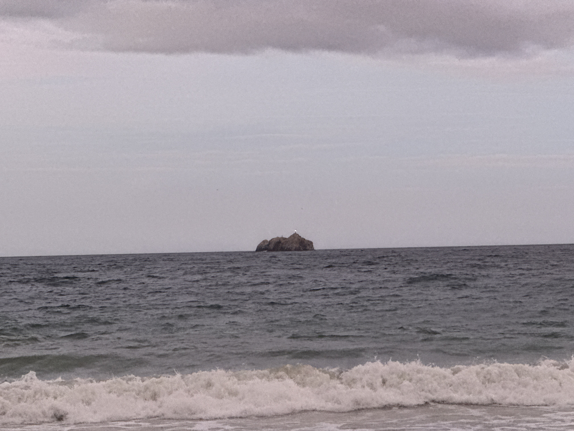 The image presents a tranquil yet dramatic coastal scene, likely captured in Porlamar, Venezuela. The frame is dominated by the sea and sky, with a clear horizon line. The sky above is overcast, featuring various shades of light grey clouds, with a darker, more defined cloud band at the very top.
In the middle distance, a prominent rocky island or sea stack emerges from the dark, choppy waters. Perched atop this rugged island is a small, bright white structure, possibly a lighthouse or a navigational beacon, which stands out distinctly against the dark rock. The ocean itself is a deep greyish-blue, characterized by numerous ripples and small waves across its surface, reflecting the overcast conditions.
In the immediate foreground, foamy white waves are seen breaking and rolling onto a narrow strip of wet sand, indicating the shoreline. The overall lighting suggests an overcast day, possibly early morning or late afternoon, creating a muted and somewhat somber atmosphere. No human activity or visible text is present, emphasizing the raw, natural beauty of the seascape.