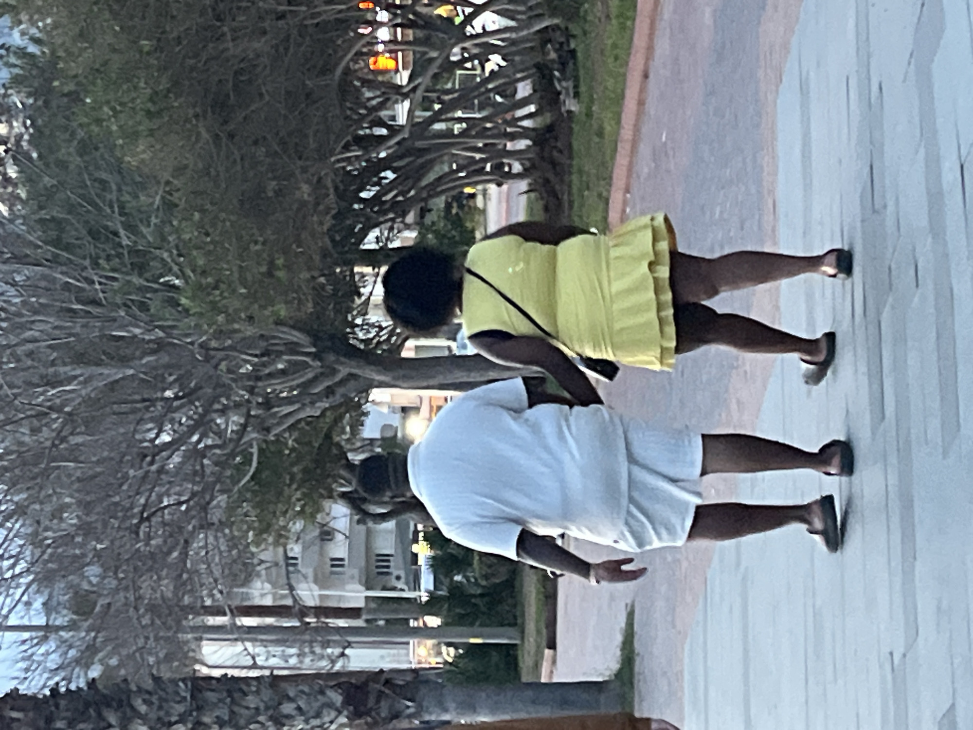 Two individuals, a man and a woman, are walking on a paved walkway in Alanya, Turkey. The woman is wearing a yellow dress and a black shoulder bag. The man is wearing a white shirt and white shorts. In the background, there are trees and buildings. The image is taken during daytime.