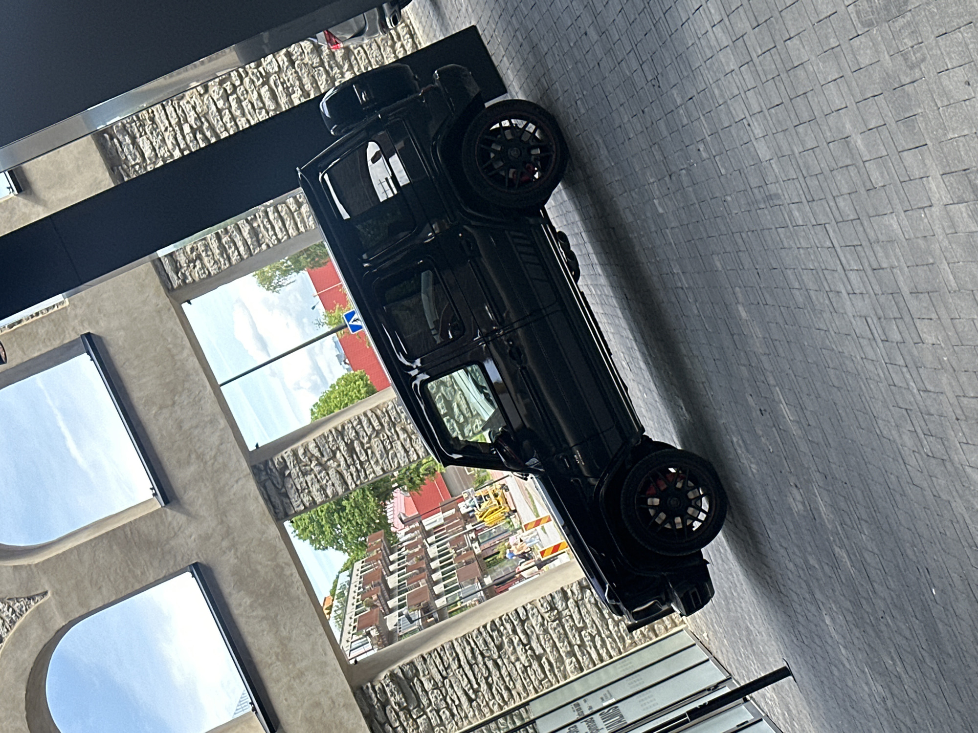 A black Mercedes-Benz G-Class SUV is parked on a cobblestone sidewalk in front of a building with arched windows and stone facade. The building appears to be a modern structure with large glass panes and dark structural elements. In the background, there is a street scene with buildings, trees, and a yellow construction vehicle. Several people are visible walking on the street. The sky is partly cloudy. The image is taken from a slightly elevated, angled perspective. The car has custom black wheels with red brake calipers. The context suggests this is likely a commercial or residential area within the city of Tallinn, Estonia, possibly near a modern development or public space.
