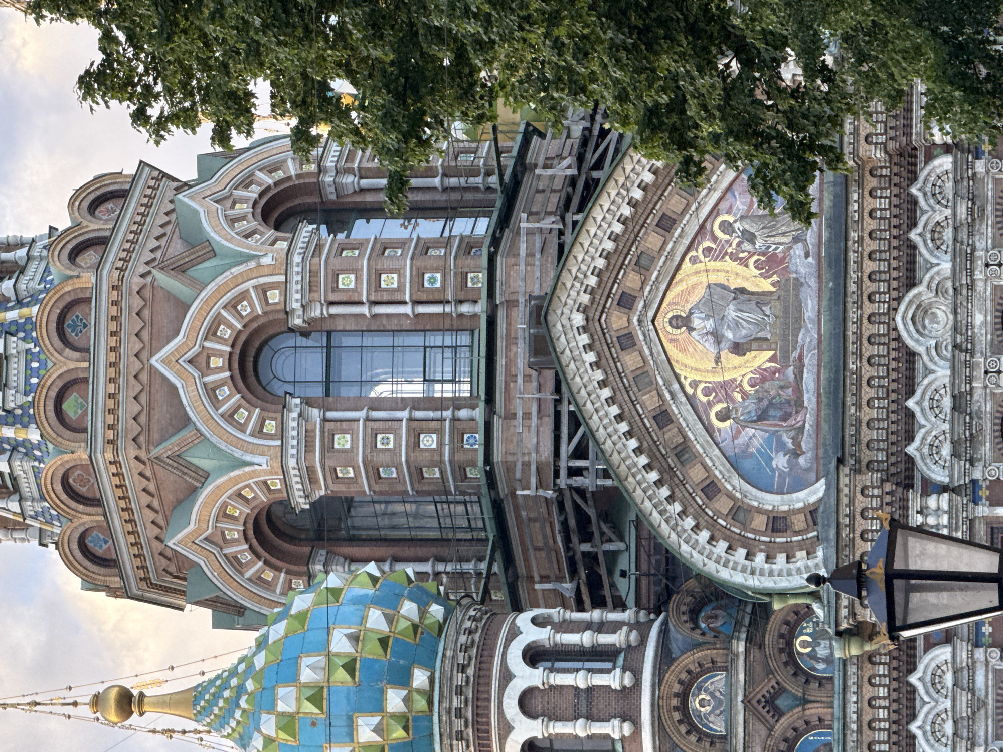 The image shows a close-up view of the exterior of the Church of the Savior on Spilled Blood in Saint Petersburg, Russia. The church is characterized by its intricate multi-colored domes and detailed mosaic decorations. In the foreground, a black lamppost with gold accents is visible. The church's facade features red brickwork, green and blue tiled domes, and a large mosaic depicting Christ enthroned with surrounding figures. Arched windows and decorative ceramic tiles are also prominent architectural elements. A large green tree with dense foliage is positioned on the right side of the frame, partially obscuring the church's structure. The sky above is overcast with diffused light. The scene captures a section of the elaborate and ornate architecture of the iconic Russian Orthodox church.