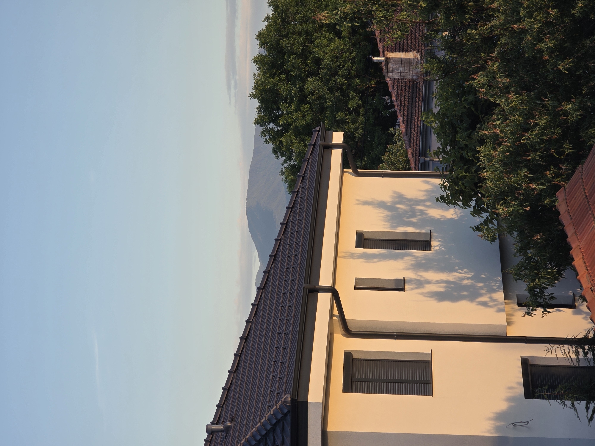 The image shows a view of a building in Yagodovo, Bulgaria. The building has a dark tile roof, cream-colored walls, and black window frames. The windows have shutters. A black rain gutter runs along the roofline. The background features a blue sky, a mountain, and green trees. Part of another building with a red tile roof is visible amidst the trees.