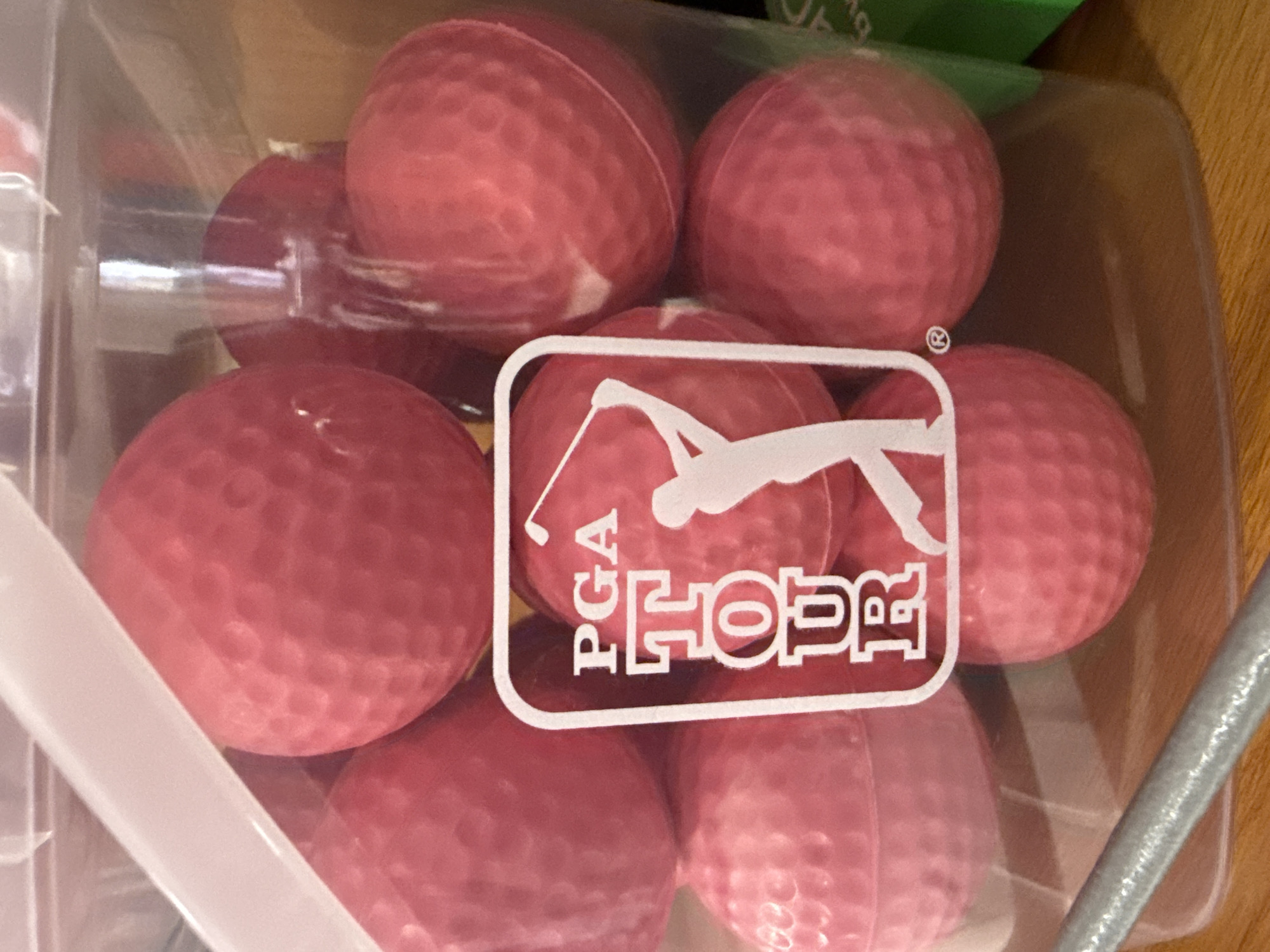 The image shows a clear plastic container filled with red golf balls. The container features a white logo with "PGA TOUR" and a silhouette of a golfer. The balls are textured with the typical dimpled surface. The photo was taken in Dania Beach, Florida, United States.