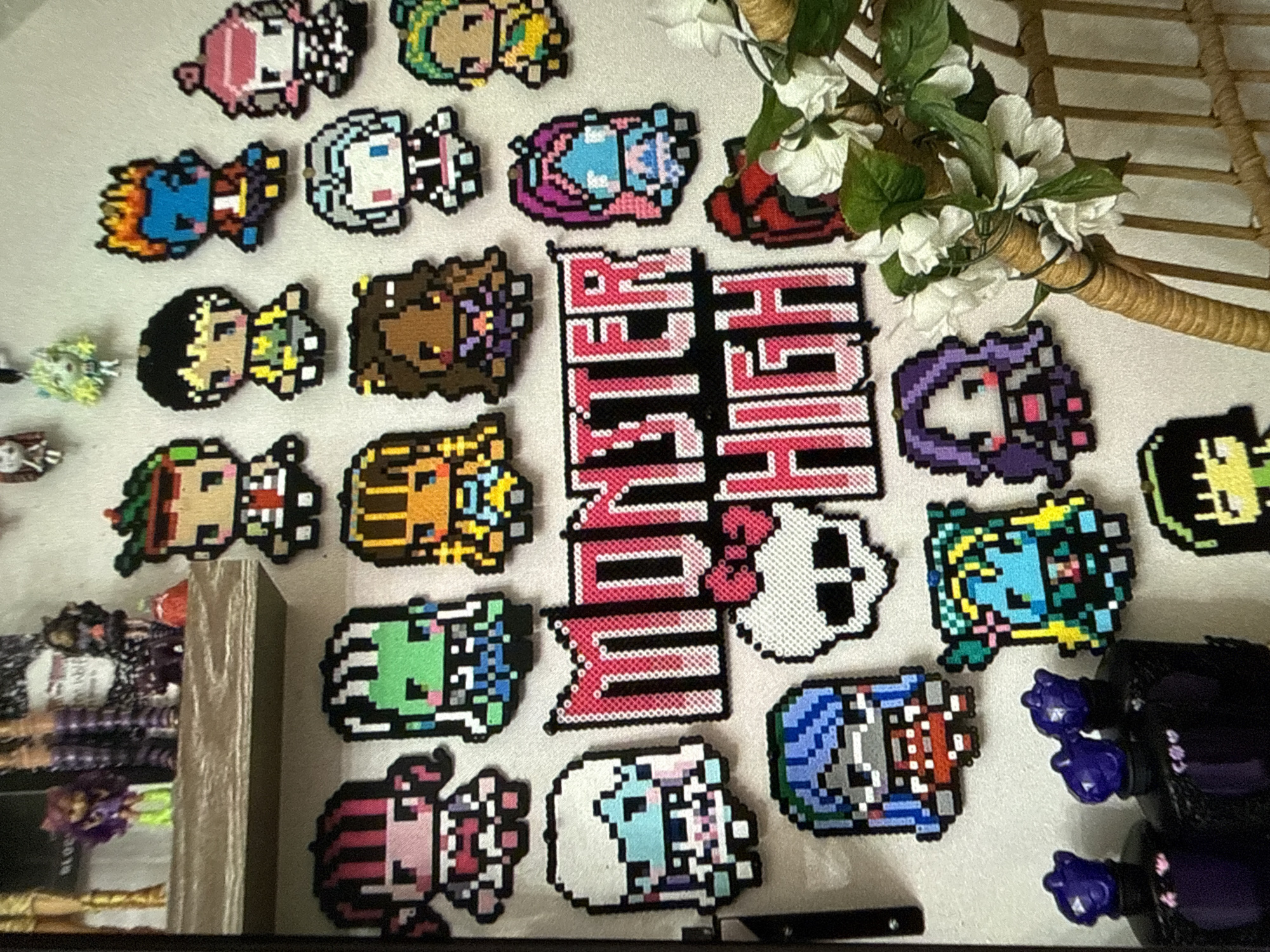 The image shows a collection of perler bead sprites arranged on a white surface. The sprites are of various characters, including several Monster High characters, indicated by the "Monster High" logo made of perler beads. The characters are depicted in a pixelated style. A floral arrangement is visible in the upper right corner of the image. Part of a display case containing dolls is visible on the left. The scene is indoors, likely in a collector's display area. The location is unknown.