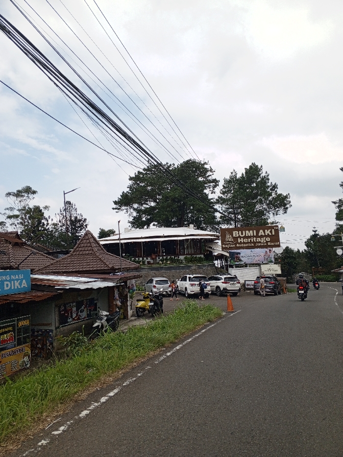 The image depicts a roadside scene in Ciloto, Indonesia, under an overcast sky. Numerous dark electrical and communication wires span across the upper left portion of the frame. An asphalt road, with a white dashed line on its left edge, curves slightly to the right, receding into the background.
On the left side of the road, a grassy verge separates the road from a series of small, rustic single-story buildings. These structures feature peaked brown tiled roofs and corrugated metal awnings. One building displays a partially visible sign that reads "RUNG NASI BU DIKA." Several motorcycles are parked near these buildings, including a yellow scooter and two dark-colored scooters.
To the right of the road, an orange traffic cone is positioned near an elevated parking area. This parking lot contains multiple parked vehicles, including several white SUVs, a silver sedan, and other dark-colored vehicles. Several individuals, consisting of both adults and children, are observed standing and moving within this parking area, some near the parked cars and others closer to an entrance.
Behind the parking lot, a large, prominent brown sign with white text identifies "BUMI AKI HERITAGE" and "SAJIAN AUTENTIK JAWA BARAT." Further back and higher in elevation, the main structure of Bumi Aki Heritage is visible: a large, circular building with a distinctive white roof and dark-paneled walls, surrounded by dense green evergreen trees.
In the mid-distance on the road, two motorcycles are present, with one actively moving away from the viewer. The background features additional evergreen trees and distant hills partially obscured by atmospheric haze. A street light pole is visible on the left side, behind the smaller buildings.