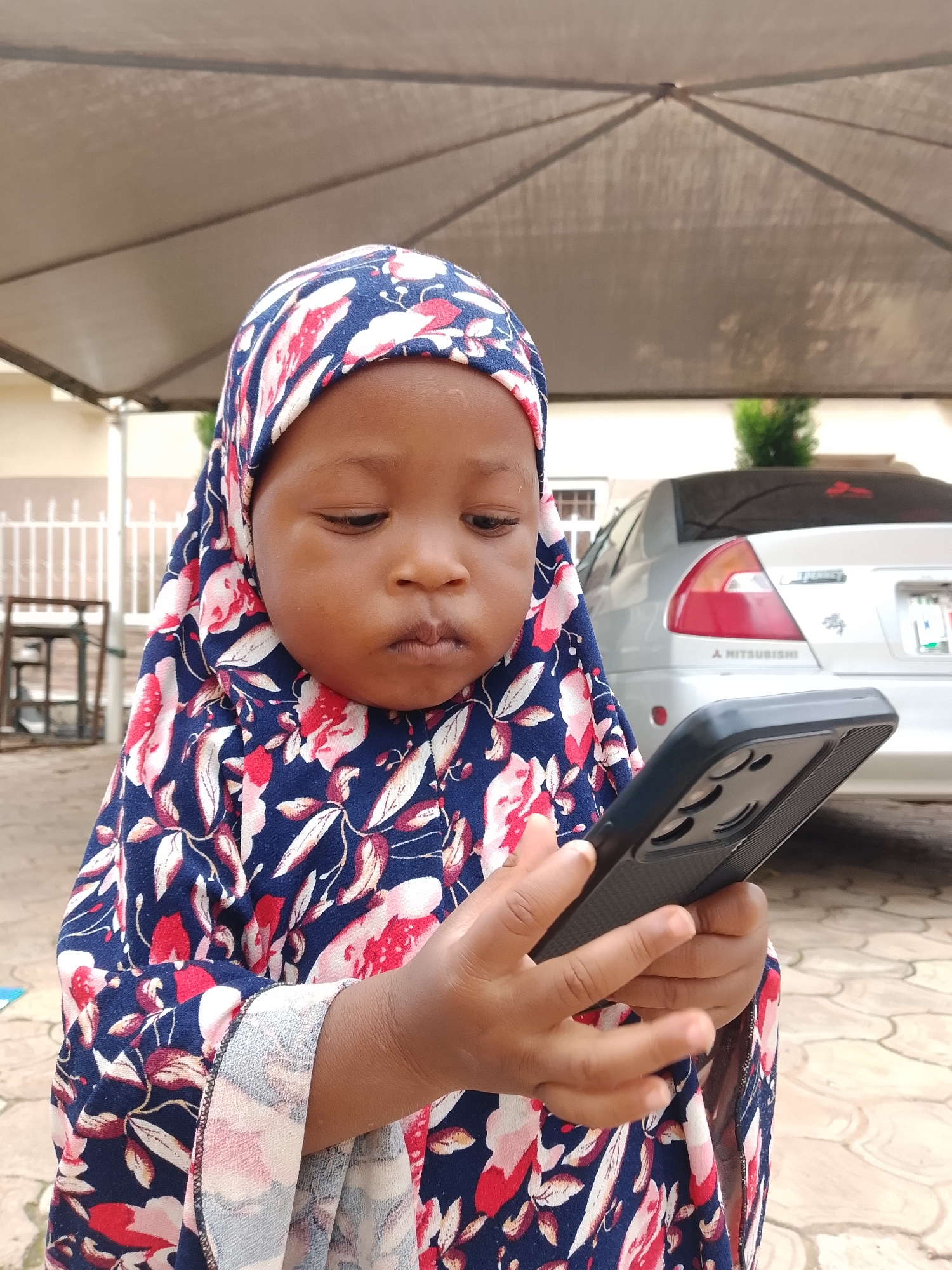A young child wearing a blue and red floral hijab and dress is holding a black smartphone. The child is looking down at the phone with a focused expression. In the background, a silver Mitsubishi sedan is parked, and behind it, a building with white railings can be seen. The scene appears to be an outdoor setting, possibly a residential area in the city of Jos, Nigeria, under a beige canopy structure. The ground is paved with interlocking stones.