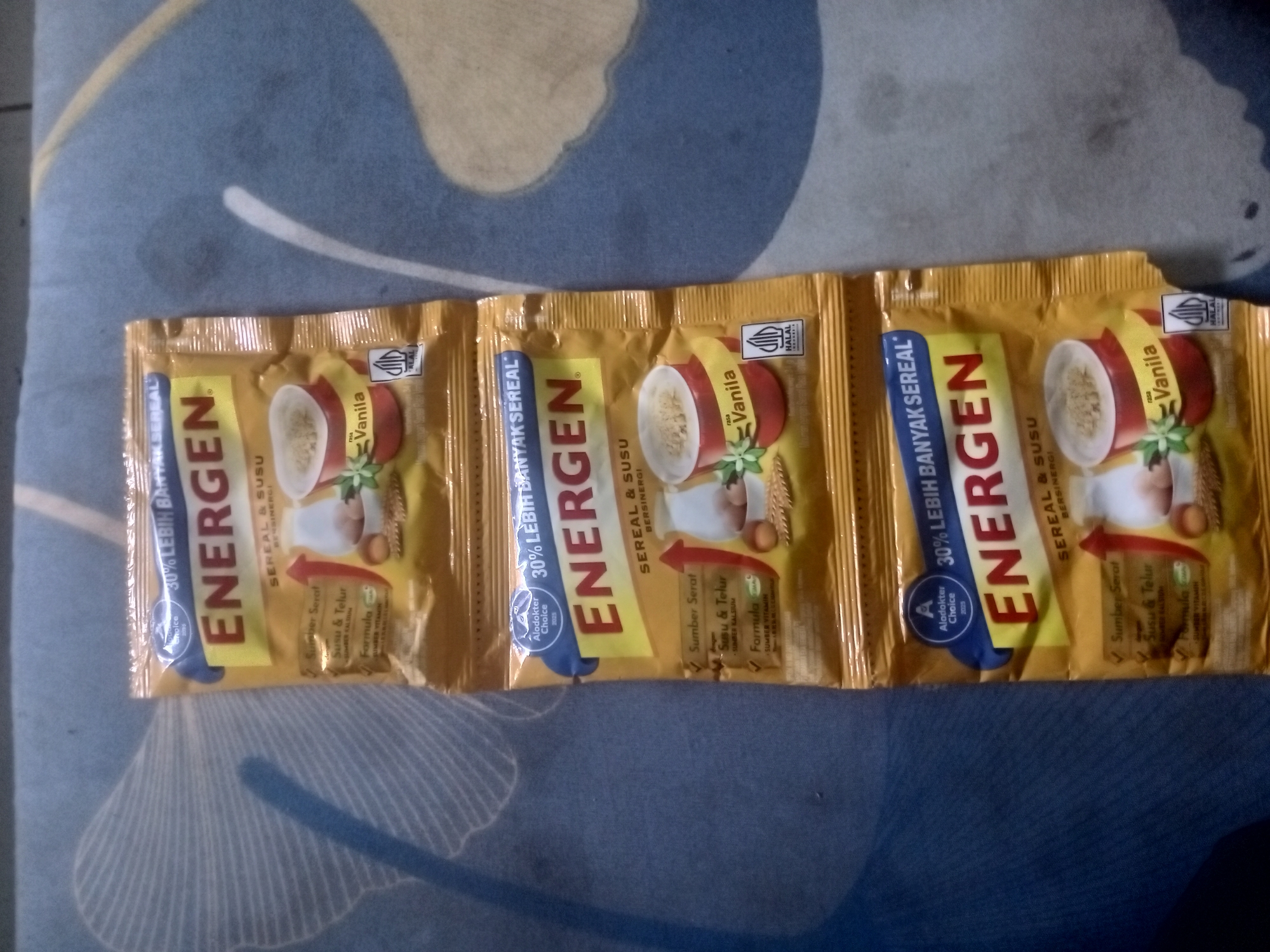 The image shows three foil packets of Energen cereal drink laid out on a blue and yellow patterned surface. The packets are golden brown with red and blue accents. Each packet has the "Energen" logo prominently displayed in large red letters. Text on the packets indicates "30% LEBIH BANYAK SEREAL" (30% MORE CEREAL) and "SEREAL & SUSU BERSINERGI" (CEREAL & MILK SYNERGISTIC). There are illustrations of a bowl of cereal, milk, and possibly eggs on each packet. The packets appear to be arranged in a row, horizontally across the frame. There is no visible context suggesting a specific location in Badas, Indonesia, or any people, actions, or interactions. The overall environment is a flat surface with a distinct pattern.