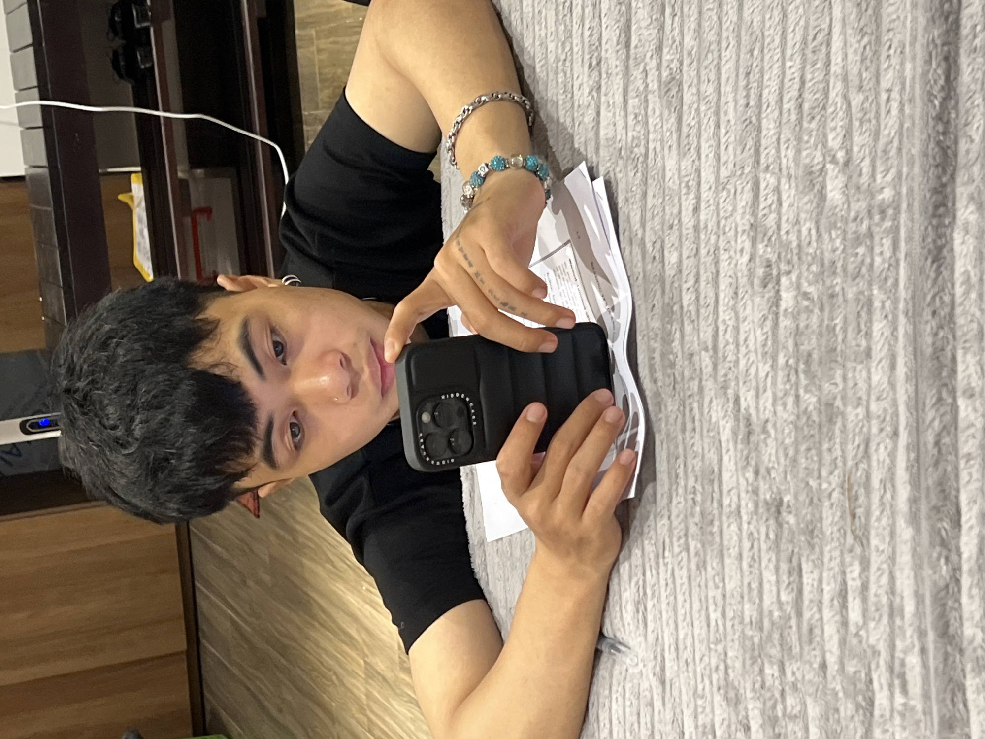 A person with dark hair and a nose piercing is holding a black phone with three camera lenses. The person is wearing a black shirt and has two bracelets on their left wrist. They are holding the phone over a pile of papers on a textured gray surface. The location is unknown and there is no indication in the image that it is Đà Nẵng, Vietnam.