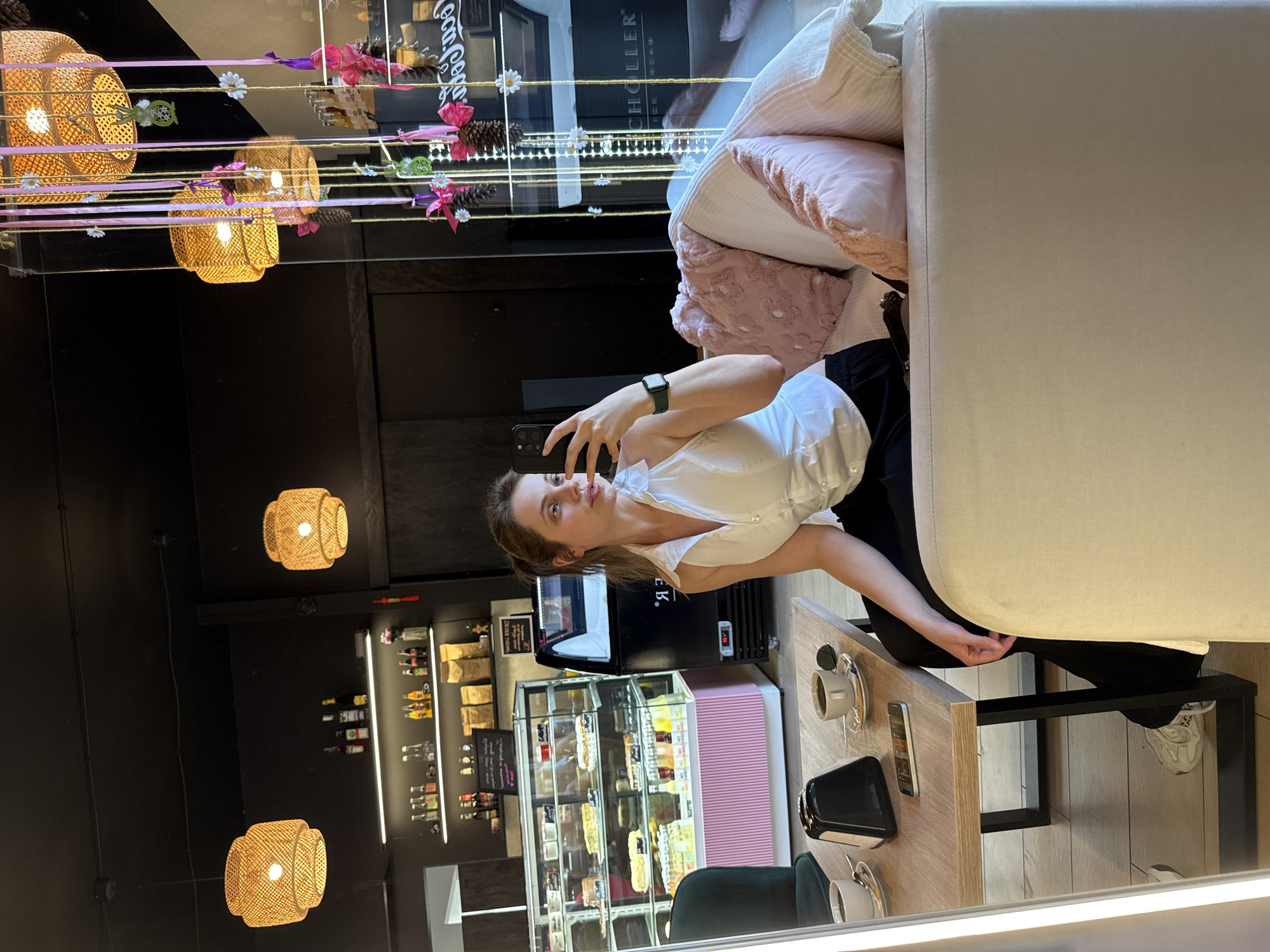 A woman is taking a selfie in a brightly lit cafe. She is wearing a white sleeveless top and black pants, with a watch on her left wrist. She is sitting on a couch, with her legs crossed, and is holding her phone in her right hand. In front of her is a wooden table with two cups of coffee, a napkin holder, and a smartphone. The cafe has a modern decor, with a display case filled with pastries and drinks, and shelves stocked with bottles and other items. Hanging from the ceiling are decorative lamps and ribbons. The overall atmosphere is casual and relaxed. The image is taken in Szklarska Poręba, Poland.