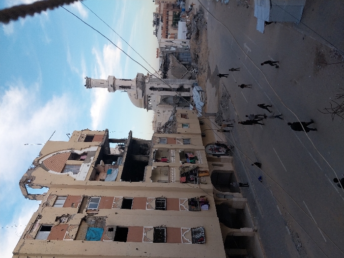 A war-torn cityscape shows a severely damaged building with gaping holes and structural collapse, next to a partially destroyed mosque with a standing minaret. A wide road runs alongside the buildings, dotted with several dark figures walking along it. Further in the distance, more buildings appear, some also showing signs of destruction. The sky is blue with scattered clouds. Overhead, power lines stretch across the scene.