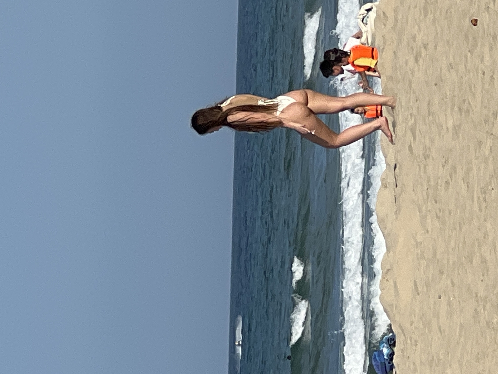 A side view shows a woman in a white bikini walking on a sandy beach with the ocean in the background. She has long, dark hair and is facing away from the camera. The ocean has waves and there are a few people in the water. In the foreground, there are two children wearing orange life vests and an adult. The scene appears to be on a beach in Da Nang, Vietnam.