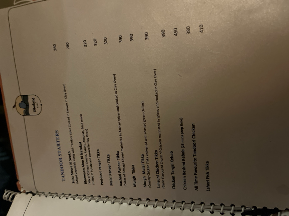 This media file displays a close-up, slightly angled view of a page from a restaurant menu, likely from "Windsong Cafe" as indicated by the logo featuring a bird or fruit icon. The page is titled "TANDOOR STARTERS" and lists various Indian appetizer dishes with their corresponding prices.
The menu items include vegetarian options like "Subz Mewa Ki Seekh" (280), "Bharwaan Aloo Ki Nazakat" (280), "Peri-Peri Paneer Tikka" (320), "Malai Paneer Tikka" (320), and "Aachari Paneer Tikka" (320). Non-vegetarian options feature chicken and fish, such as "Murgh Tikka" (390), "Murgh Malai Tikka" (390), "Lahsuni Chicken Tikka" (390), "Chicken Tangri Kebab" (390), "Chicken Reshmi Kebab" (450), "All Time Favourite Tandoori Chicken" (380), and "Lahori Fish Tikka" (410). Some items have brief descriptions, for example, "Chicken Reshmi Kebab" notes a "25 mins prep time".
The menu page has a cream-colored background with black text. A metal spiral binding is visible along the left edge, indicating a bound menu. The lighting appears warm, suggesting an indoor setting. The image provides a factual glimpse into the culinary offerings of a cafe in Dibrugarh, India.
