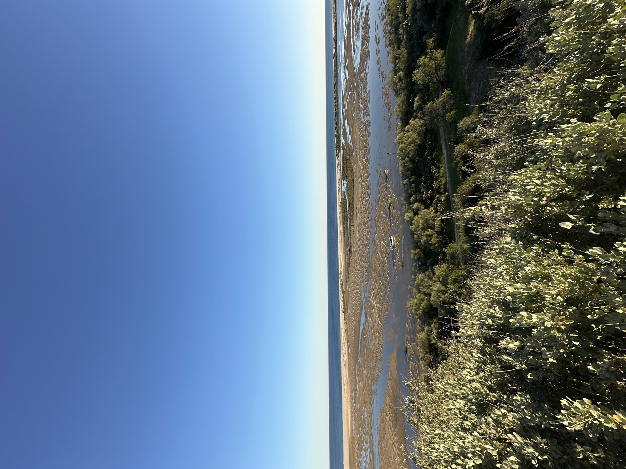 The image shows a beach scene in Vila Real de Santo António, Portugal. The sky is clear and blue. To the right of the sky is the shore with water and green vegetation and trees.