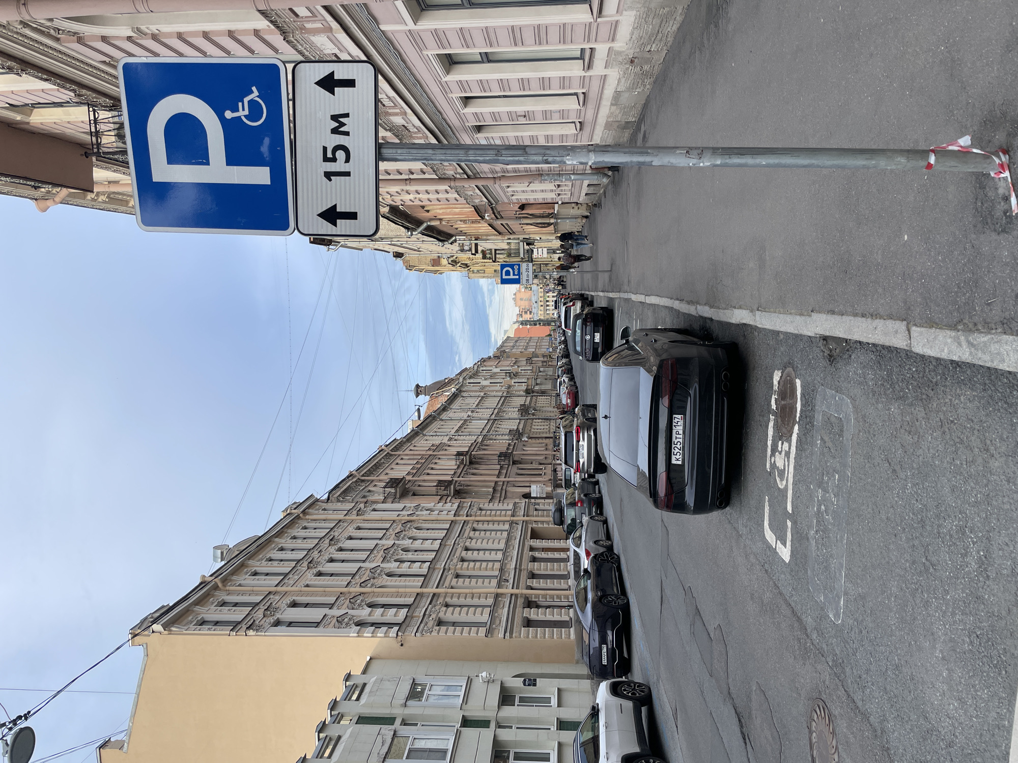 The image presents a street scene in Saint Petersburg, Russia, captured with a tilted perspective, showing the street and buildings on a vertical plane. Ornate, multi-story historical buildings with numerous windows and decorative facades, predominantly in light beige and pinkish tones, line both sides of the street, stretching into the distance.
In the foreground, a blue parking sign with a white 'P' and a wheelchair symbol denotes accessible parking. Beneath it, a rectangular sign specifies "15M" (15 meters) with upward and downward arrows, indicating the length of the designated zone. This sign is affixed to a metal pole, which has red and white striped warning tape wrapped around its base. Further down the street, another blue parking sign is visible, possibly displaying hours like "08-20:00."
Numerous cars are parked bumper-to-bumper along the right side of the asphalt street. The black car closest to the viewer has a Russian license plate "K525TP 142." White painted markings, including what appears to be "L7" or similar symbols, are visible on the road surface, along with manhole covers. A few pedestrians can be seen walking on the sidewalk in the distance.
Above the historic architecture, a clear to partly cloudy blue sky is visible, traversed by overhead wires. The scene depicts a typical daytime urban environment, highlighting the blend of historical architecture with contemporary street infrastructure and daily city life.