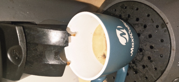 A dark-colored capsule coffee machine is dispensing a hot beverage into a teal mug on a black, perforated drip tray, likely within a domestic or commercial indoor setting in Vendin-le-Vieil, France. Two streams of light brown liquid with visible steam or vapor are actively flowing from the machine's twin spouts into the mug. The mug, which has a white interior, contains a small volume of coffee with a light brown crema layer and some darker residue. A white logo, consisting of a stylized "M" above the word "MaxiCoffee," is prominently displayed on the side of the mug. The drip tray exhibits dark liquid splashes and residue. The upper left side of the coffee machine features a circular depression with an embossed design.