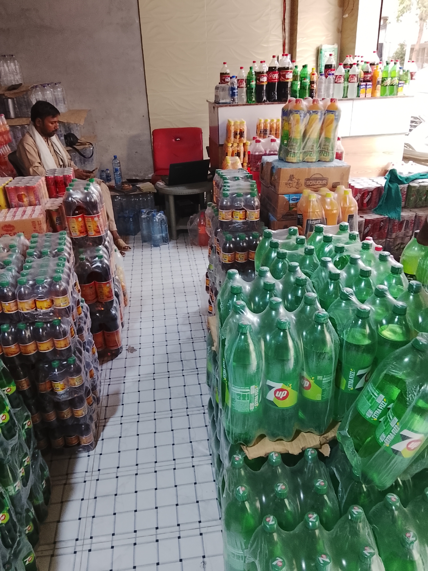 Captured indoors at what appears to be a retail space, possibly in Gulgasht Colony, Pakistan, a man is seated to the left, engaged with his mobile phone. He is surrounded by stacks of bottled beverages, including water, soda, and juice, which fill much of the foreground and extend into the background. A red chair and a laptop on a small table are situated near the man, suggesting an office setup within the store. Shelves along the back wall display an assortment of additional bottled drinks. The floor is tiled, providing a grid-like pattern throughout the scene. The scene is well-lit, though with a slightly overcast feel.