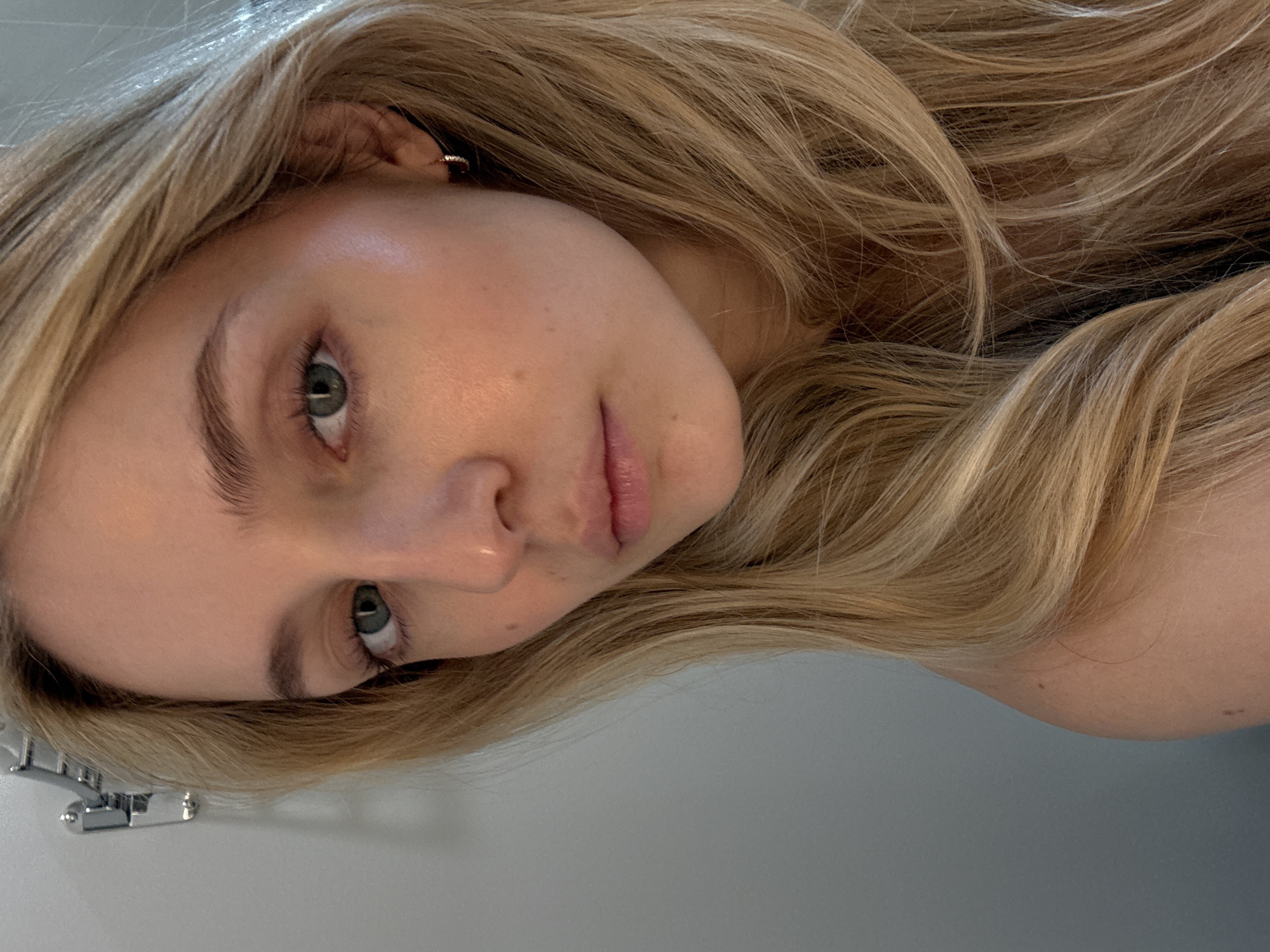 A close-up, head-and-shoulders, profile shot of a young Caucasian woman with long, wavy, blonde hair. Her head is tilted slightly to her left, and her gaze is directed downwards and to her left. She has light skin with a few freckles visible on her cheek. Her eyes are a greenish-blue hue, and she has light brown eyebrows. Her lips are full and appear to have a slight sheen. She is wearing a small, gold hoop earring in her visible ear. The background is a plain, light gray wall. There is a metallic towel rack visible on the left side of the image, mounted on the wall. The lighting appears to be soft and natural, casting a gentle glow on her face. The overall impression is of a candid, casual portrait. The image was taken in Miami, United States of America.