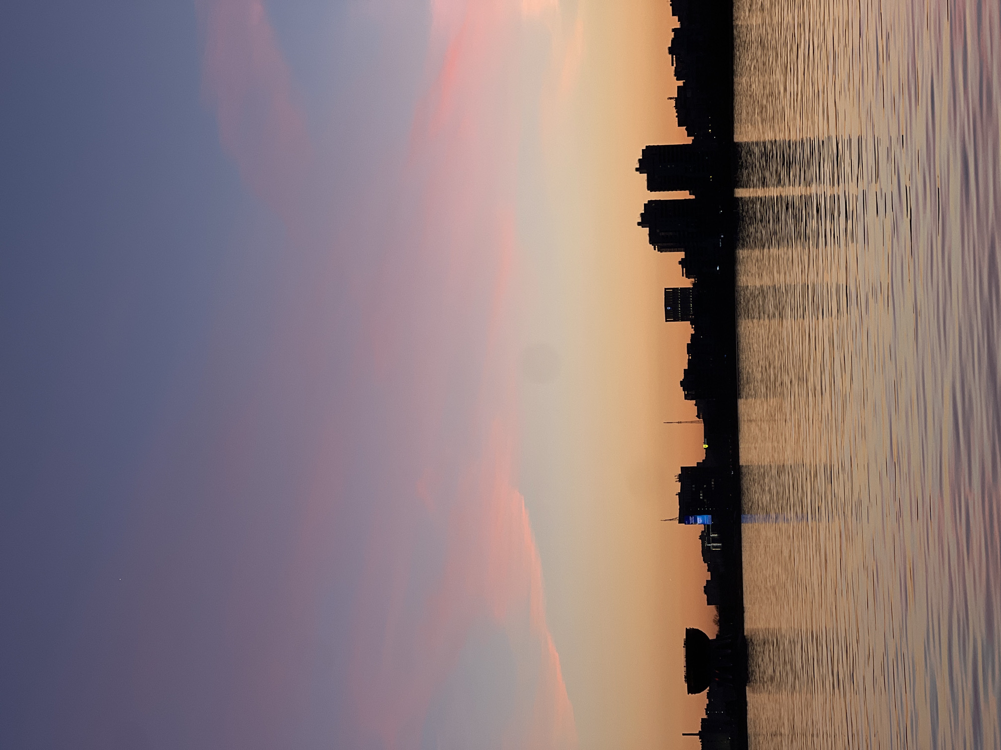 The image presents a serene panoramic view of the Kazan city skyline in Russia, captured during either dusk or dawn. The sky, which occupies the upper portion of the frame, displays a striking gradient, transitioning from deep blue and purple on the left to warm pink and orange hues towards the right. Scattered, soft clouds are visible, particularly in the pinker sections of the sky.
A dark silhouette of the city stretches horizontally across the middle, showcasing numerous buildings of various heights. A prominent feature is a building with a bright blue-lit facade, displaying indistinct white text, located roughly in the center-right of the skyline. To the left, a distinct dome-shaped structure or monument is visible among the other buildings.
Below the cityscape, a body of water, likely a river or lake, fills the lower half of the image. The water's surface is textured with ripples, reflecting the vibrant colors of the sky and the dark silhouettes of the city buildings, creating a sense of depth and tranquility. No people are visible, emphasizing the expansive and calm urban landscape. The overall mood is peaceful and picturesque, highlighting the beauty of the city at twilight.
