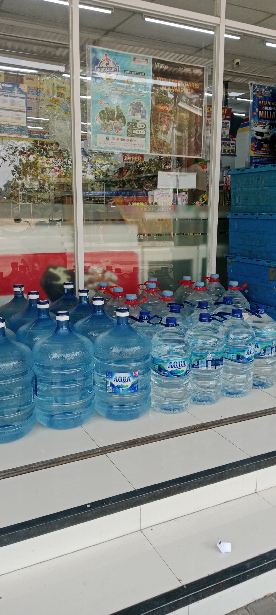 The image shows a storefront, likely in Maguwoharjo, Indonesia, featuring a display of large bottled water jugs lined up on a tiled floor in front of glass doors. The jugs are of the Aqua brand, and some have blue handles and red caps, while others have blue caps. Behind the jugs, through the glass, shelves stocked with various products are visible, along with promotional posters advertising Indomaret and Nescafe. There are also blue plastic crates stacked to the right of the storefront. The environment suggests a retail setting, possibly a convenience store or minimarket, with a focus on beverage sales. The lighting indicates it is daytime.
