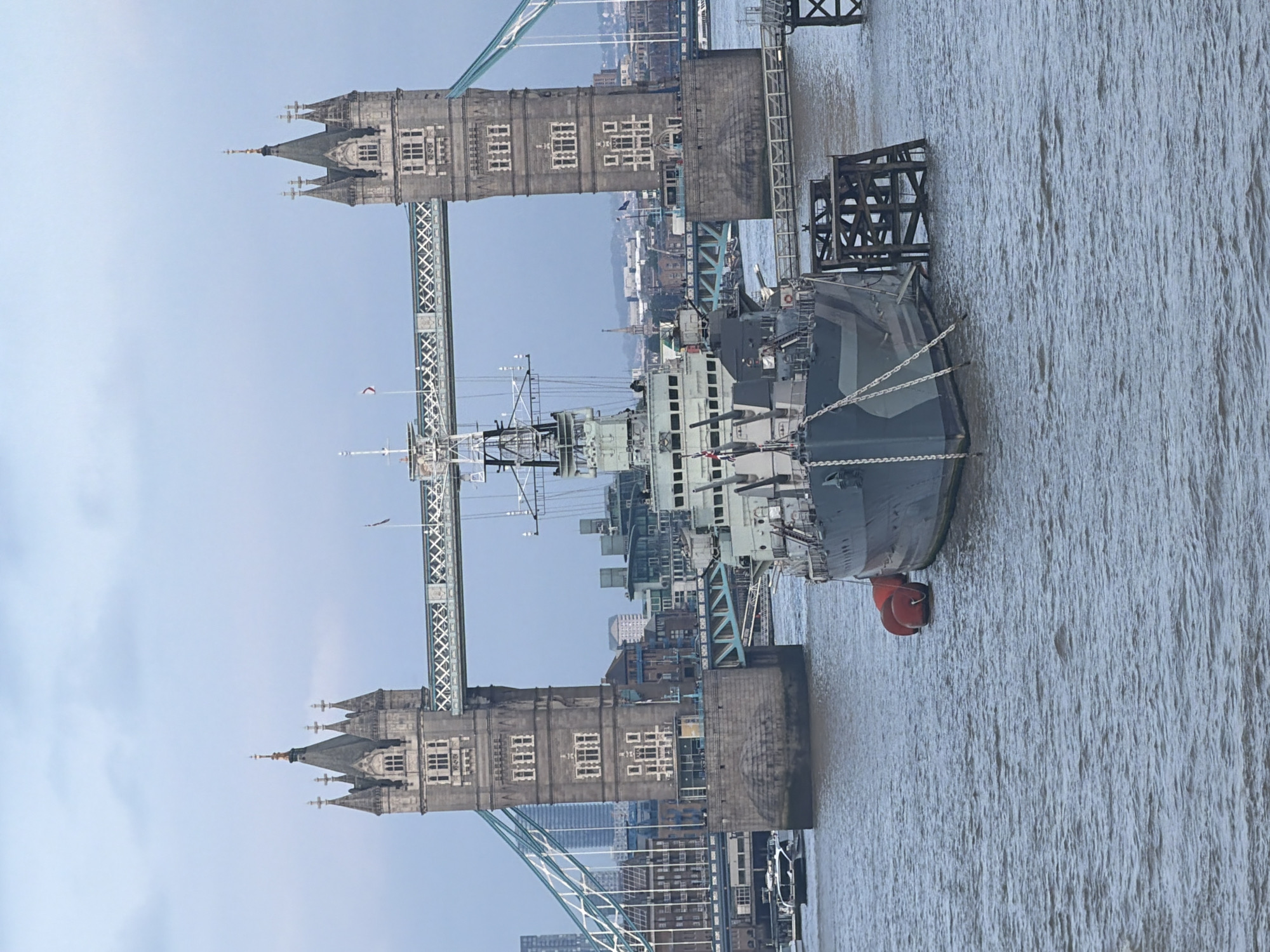 The image captures a view of the Tower Bridge in London, UK. The Tower Bridge is a combined bascule and suspension bridge in London, built between 1886 and 1894. In front of the bridge, a Royal Navy warship is berthed, showcasing its complex superstructure. The river's surface is rippled, reflecting the overcast sky above. The scene portrays an iconic blend of London's historic architecture and maritime heritage.