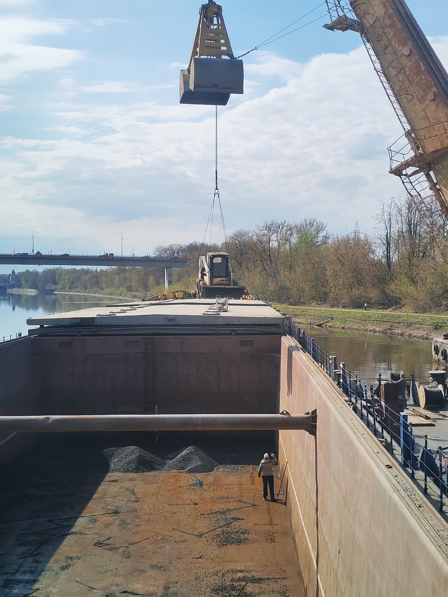 The image shows a barge being loaded with gravel by a crane. The barge is on a canal in Yakhroma, Russia. In the foreground, a worker with a broom is standing in the barge's hold, near a pile of gravel. Above the barge is a crane with its bucket suspended in the air. On the barge's deck sits a construction vehicle. The canal is lined with trees, and a bridge is visible in the background.