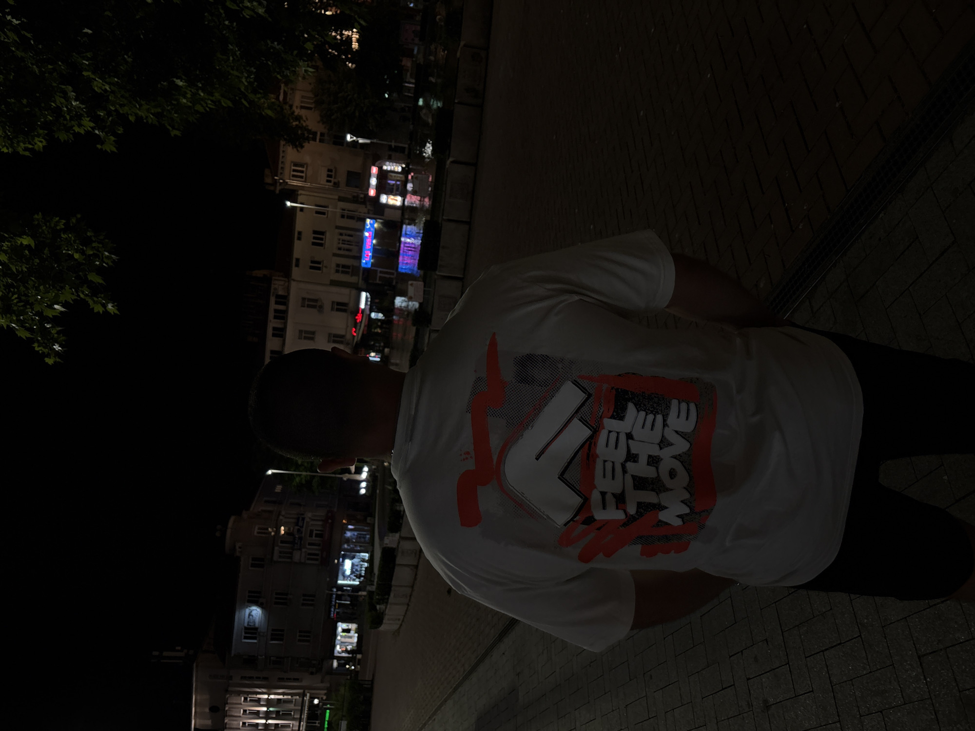 An adult male subject is observed from behind at night in an urban pedestrian area, specifically Nezavisimost Square (Opera Square) in Varna, Bulgaria. The subject wears a white, short-sleeved t-shirt with a prominent graphic on the back. The graphic features a stylized, angular white "F" outlined in black, superimposed over a background of red and dark grey/black distressed patterns. Below this, the phrase "FEEL THE MOVE" is displayed in bold, white, block letters within a red, rectangular, distressed border. The subject also wears dark shorts.
The environment comprises multiple multi-story buildings in the background, characteristic of European urban architecture, with various shops and businesses occupying the ground floors, identifiable by their illuminated signage in red, blue, and white. Overhead, the sky is dark. Illumination is provided by streetlights, visible as points of light and glowing reflections, and the light emanating from commercial establishments. The ground is paved with dark, rectangular blocks arranged in a pattern typical of pedestrian zones. Low, rectangular concrete planters or barriers separate different sections of the square. Dark foliage from trees is visible in the upper sections of the image. Reflections of light on portions of the pavement suggest a wet surface or the presence of nearby water features.