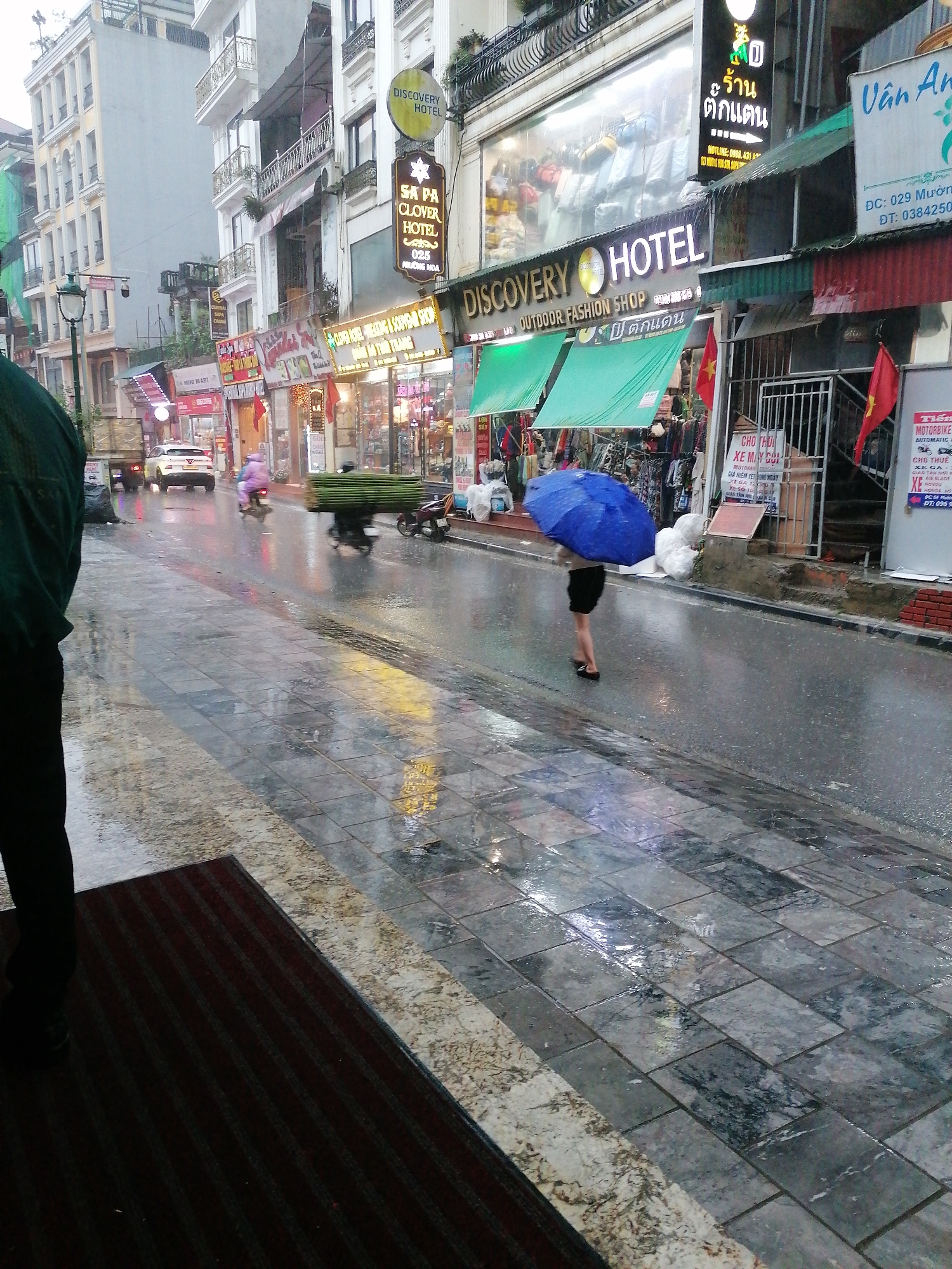 The image shows a wet street scene in Sa Pa Ward, Vietnam. It is raining, and the cobblestone street is reflecting light. In the foreground, a person's leg and part of their body are visible on the left edge of the frame. In the middle ground, a woman with a blue umbrella is walking along the street, away from the viewer. Further down the street, there are various shops and businesses with signs in both Vietnamese and English, including "Discovery Hotel" and "Sa Pa Clover Hotel". Motorbikes are seen parked and moving in the street. The buildings are a mix of old and new architecture.