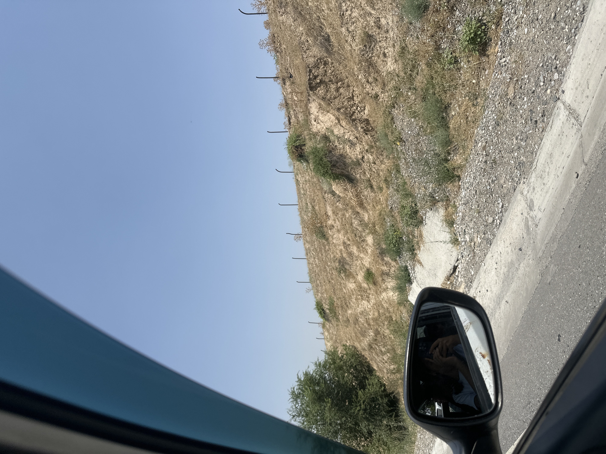 The image shows a view from inside a car looking out the side window. A side-view mirror reflects a person inside the car, holding a smartphone. Outside, a dry, grassy embankment slopes upwards, lined with a fence of dark posts. Green trees are visible on the left side of the embankment, and a clear blue sky fills the upper portion of the frame. The road in the foreground is paved, with a concrete gutter on the right. The scene appears to be a roadside in a rural or semi-rural area, consistent with the outskirts of Suzak, Kyrgyzstan, given the dry vegetation and open landscape.