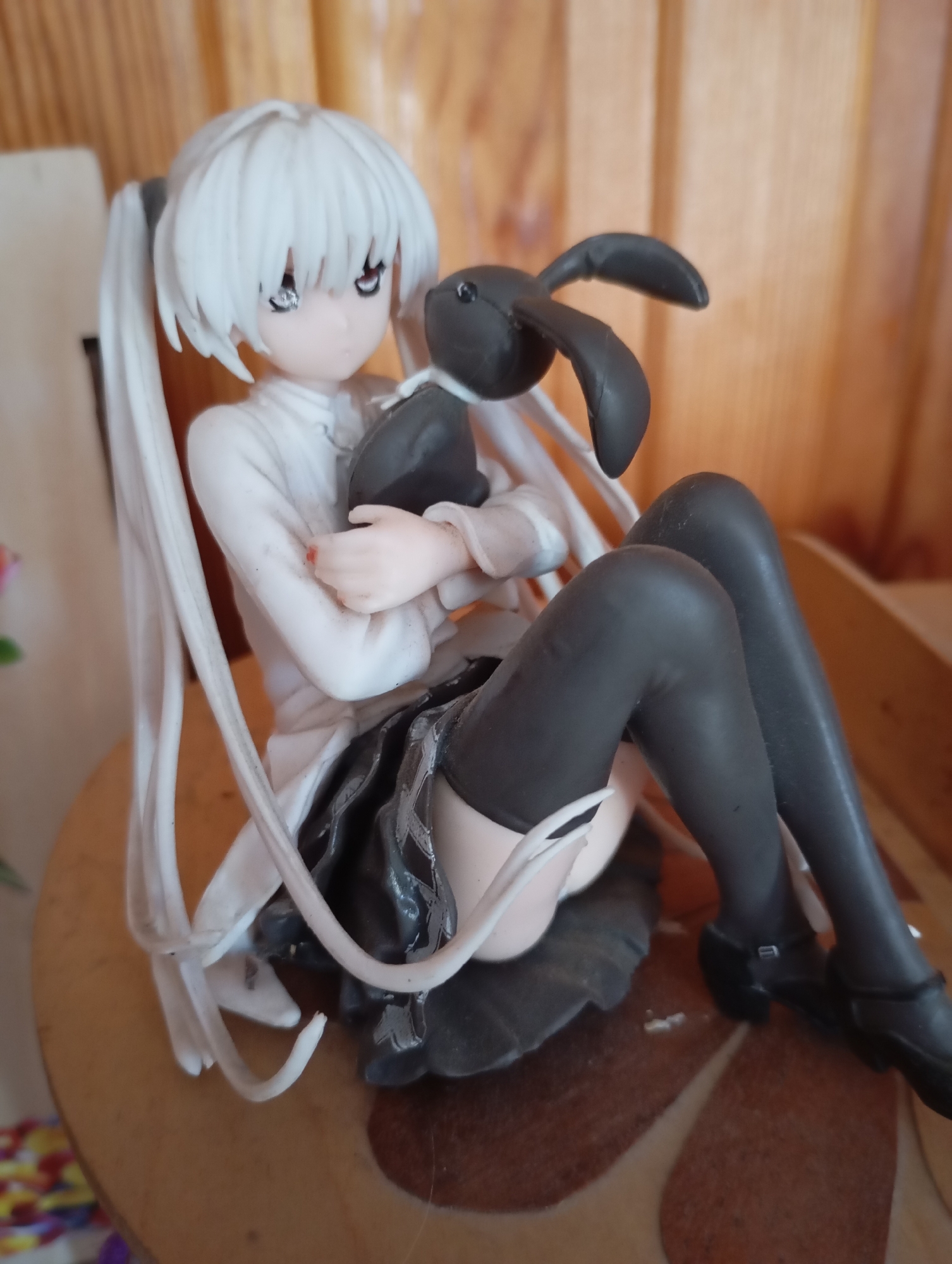 A figurine of a young woman with white hair styled in pigtails is seated, clutching a black plush rabbit. The figurine wears a white collared shirt, a dark pleated skirt, and dark stockings. It is placed on a round wooden surface with a patterned inlay. In the background, a wooden wall with vertical planks is visible. The location is specified as Lvoviha, Russia.