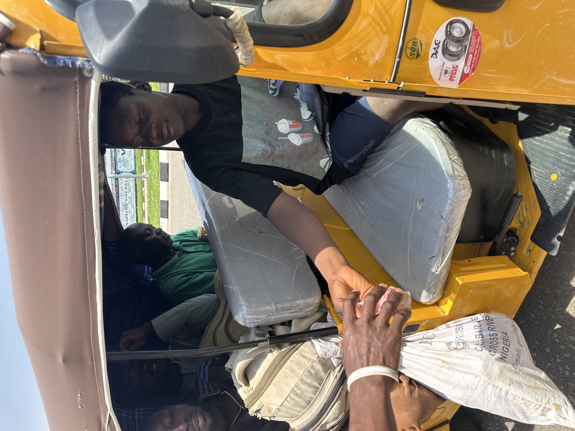 In the city of Abuja, Nigeria, a bright yellow auto-rickshaw is parked on a paved surface. The vehicle carries several passengers. The driver, a young Black male with short hair, is wearing a black t-shirt with a graphic print and dark blue shorts. He is seated and looking forward. In the passenger area, two other Black males are visible. One, wearing a green polo shirt, is looking down. Another, wearing a dark shirt with blue stripes, is partially obscured, with his fist raised. A third passenger is visible in the background, head down. A light-colored woven bag is being handed to a person outside the vehicle, whose hands are visible in the foreground, wearing a white wristband. The auto-rickshaw's interior is covered with plastic sheeting on the seats, and a brown fabric canopy extends over the top. Several bags are stowed in the passenger area. A sticker on the side of the auto-rickshaw displays "SON" and an oval sticker with "DAC" and "MRF TYRES" branding. In the background, a sign for "VINTAGE VILLAS" is visible, suggesting a residential development.