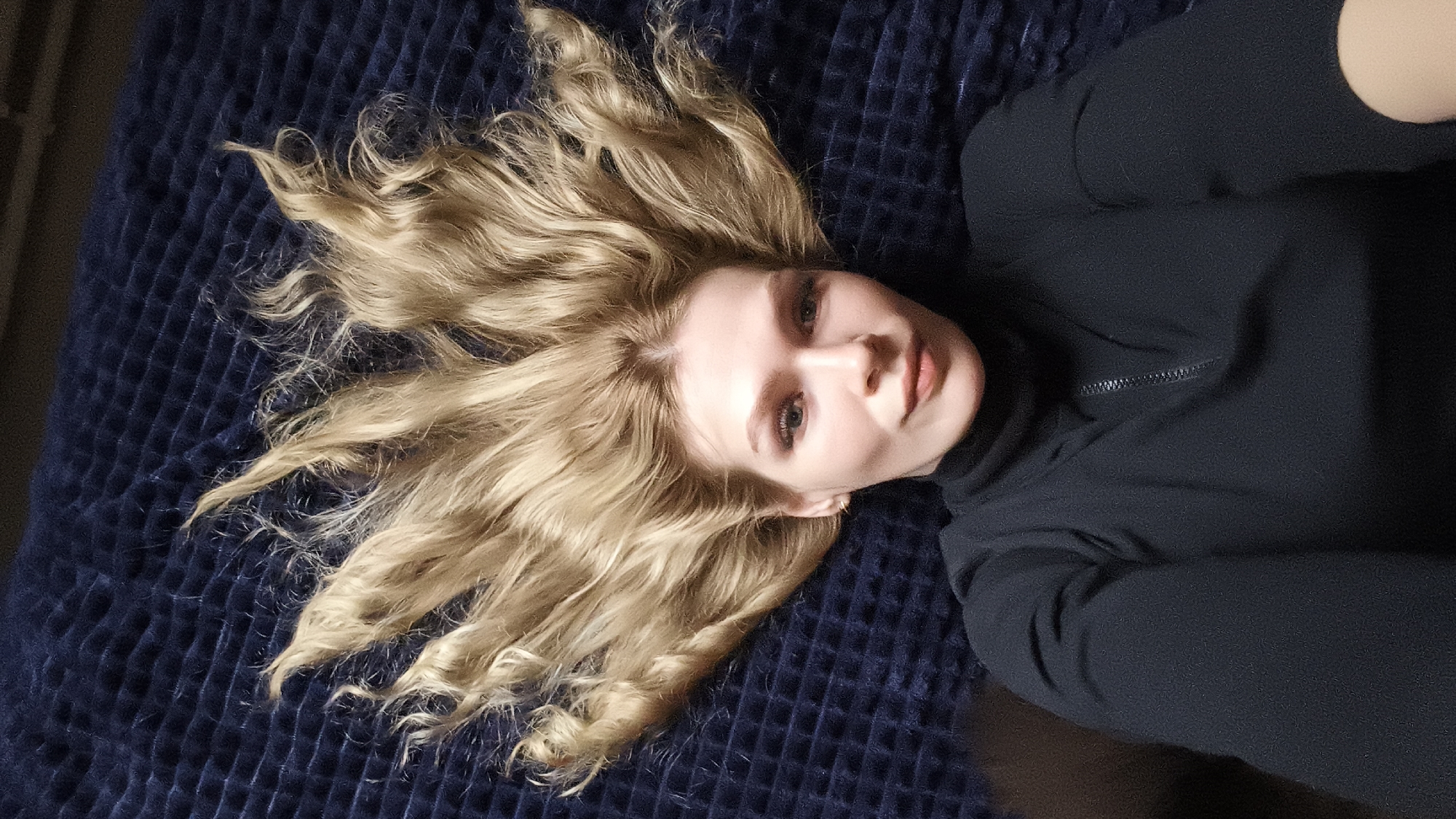 A young woman with long, wavy blonde hair is depicted lying down against a dark blue, textured background, likely a blanket or bedspread. The image is oriented such that her head is towards the upper left, with her hair fanned out around it. She has fair skin, light-colored eyes, and is looking directly at the camera with a neutral yet engaging expression. She is wearing a dark, long-sleeved top or jacket, possibly black, featuring a visible zipper detail on the chest. The lighting appears to be soft indoor light, highlighting her features and the texture of her hair. No specific activity is taking place other than posing for the camera. There is no visible text. The location context for this media file is Русский Вожой, Russia.