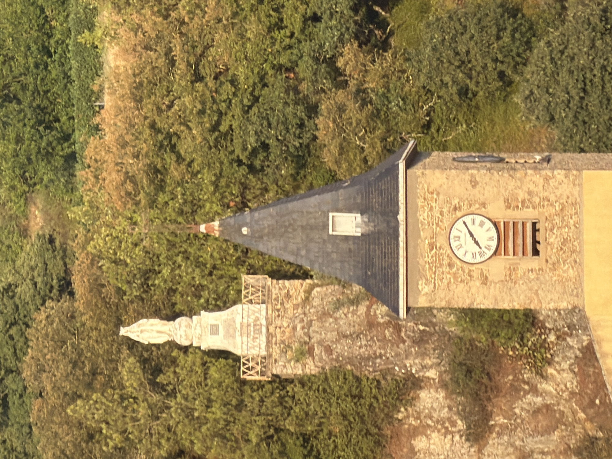 The image displays a close-up view of a church tower and a separate stone structure topped with a statue, set against a backdrop of dense green foliage. The church tower, constructed from light-colored stone, features a dark, pitched roof with a small rectangular window. Below the roofline, a circular clock face with Roman numerals is prominently displayed. A horizontal slit window is visible beneath the clock. To the left of the tower, a tall, narrow stone structure rises from a rocky cliff. This structure is topped with a decorative railing and a pedestal supporting a white statue of a robed figure, likely religious in nature. The environment is lush and green, suggesting a natural or rural setting, with trees and vegetation dominating the background. The lighting indicates it may be late afternoon or early morning, with warm tones illuminating the scene. The image is a photograph of architectural elements and natural scenery. The structures are located in Serves-sur-Rhône, France.