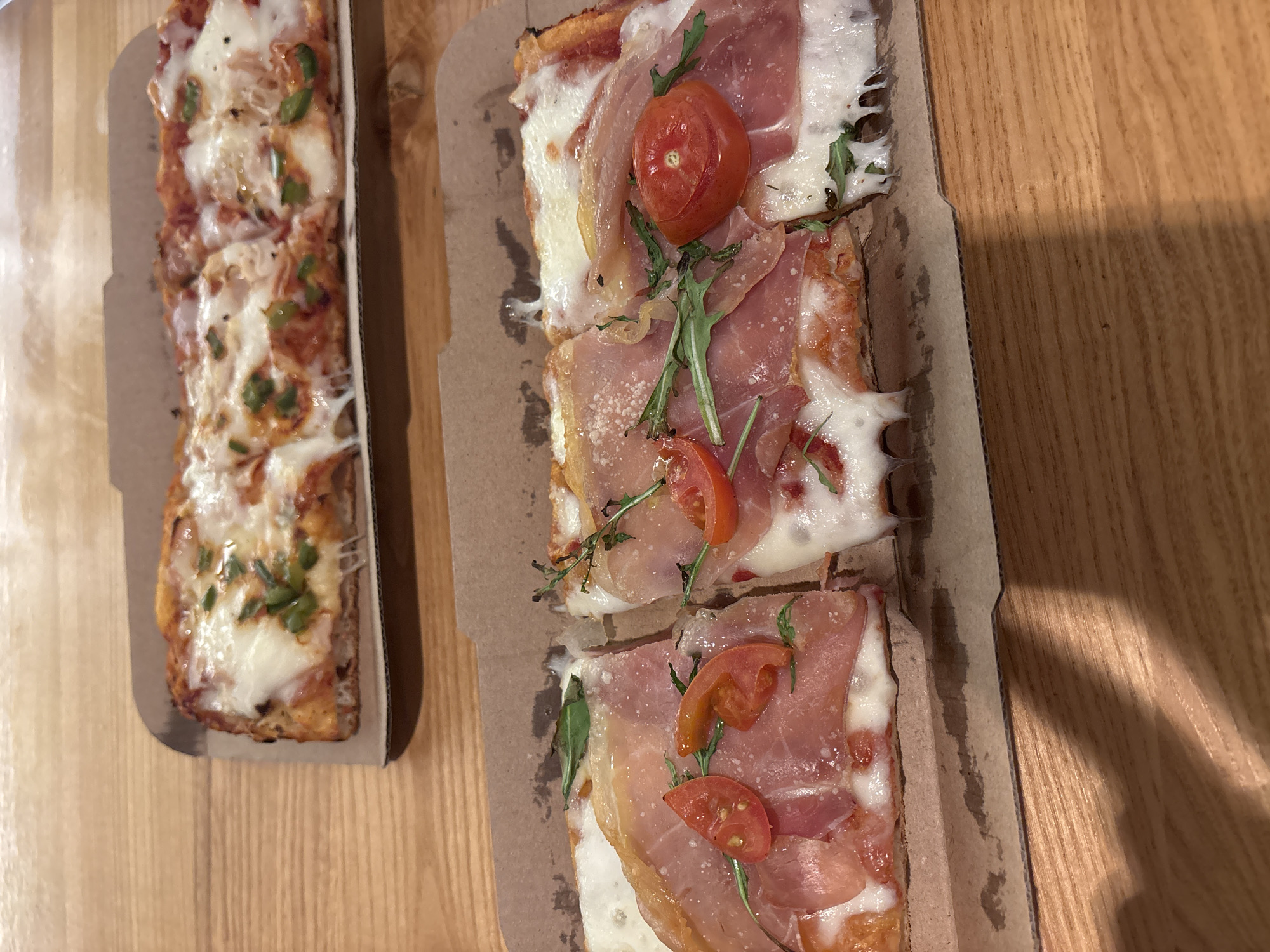 The image shows two rectangular pizzas in cardboard takeaway boxes on a light-colored wooden surface, likely a table.
The pizza on the left appears to have a tomato sauce base, mozzarella cheese, and a scattering of green toppings. The pizza on the right has a similar rectangular shape and appears to be topped with mozzarella cheese, prosciutto slices, halved cherry tomatoes, and a few green leaves.
The scene gives no specific indicators of being in Sofia, Bulgaria.