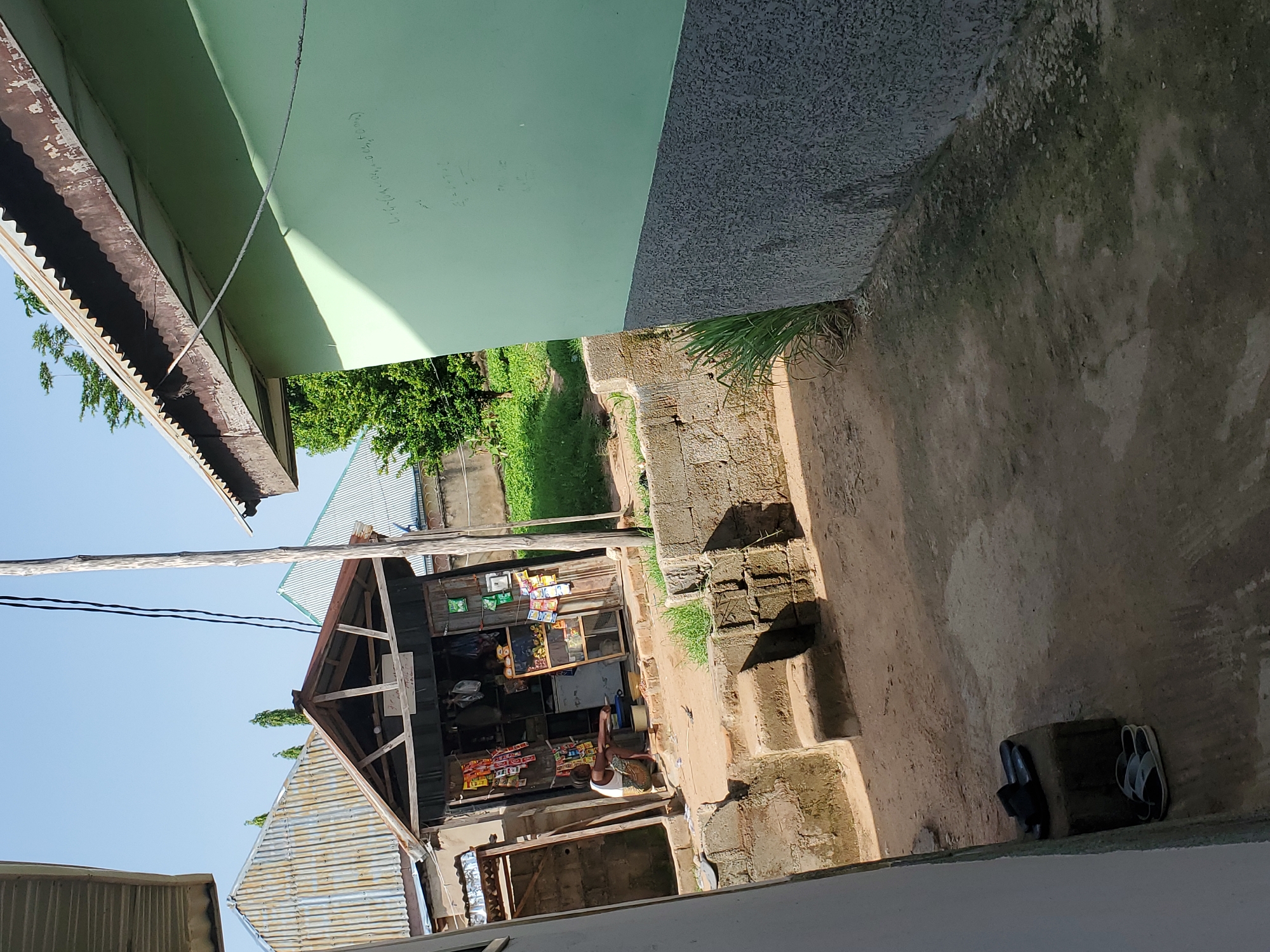 This image depicts a street-level view of a small outdoor shop in Jalingo, Nigeria. The shop, constructed with wooden beams and a corrugated metal roof, displays various goods on shelves and racks. Items such as packaged snacks and beverages are visible. A person, dressed in a white tank top and patterned shorts, is seated near the shop, appearing to be engaged in a task or resting. The scene is set in an open, earthy area, with a concrete structure and steps in the foreground. A wall, painted a light green on one side and a dark textured material on the other, runs along the upper portion of the image. The sky is clear and blue, suggesting it is daytime, likely morning or afternoon, with pleasant weather. In the background, lush green trees and foliage indicate a rural or semi-rural environment. The overall impression is one of a typical, everyday scene in a Nigerian community. There are no visible signs with text.