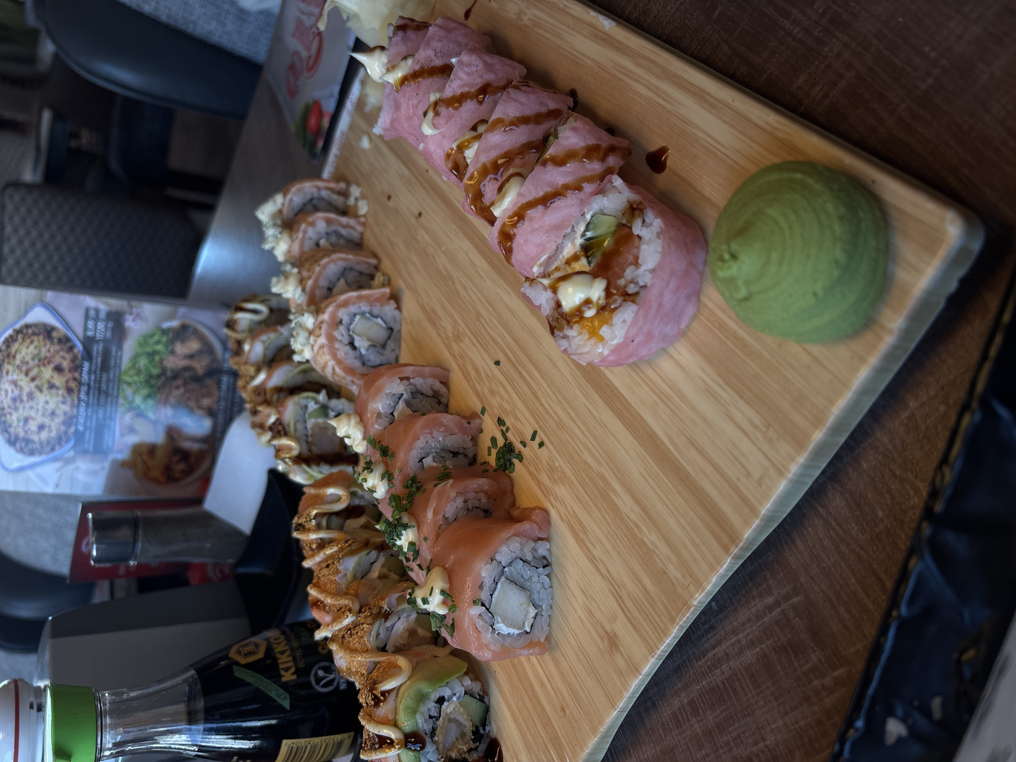 The image shows a wooden serving board containing three different types of sushi rolls and a dollop of wasabi. The rolls are arranged in three rows, with a row of sushi wrapped in a light pink ingredient at the top, followed by a row of sushi wrapped in salmon, and a bottom row featuring fried toppings. A bottle of Kikkoman soy sauce and restaurant menus are visible to the left of the serving board. The photograph appears to have been taken indoors at a restaurant, likely in the city of Varna, Bulgaria, based on the image context.