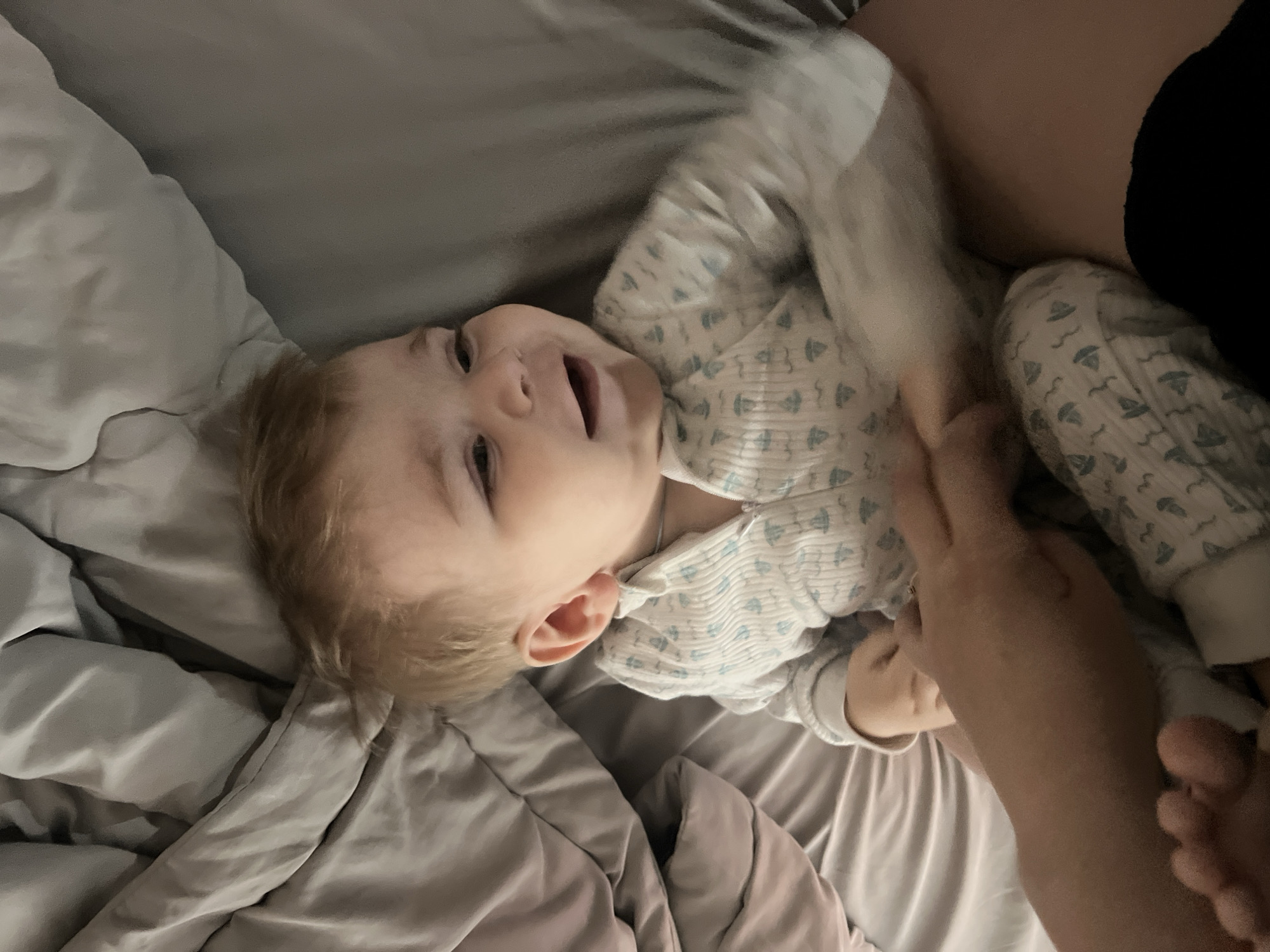 A baby wearing a white onesie with a light blue pattern is lying on a gray bed. An adult hand is near the baby. The location is not identifiable as Noviy, Russia from the provided image.