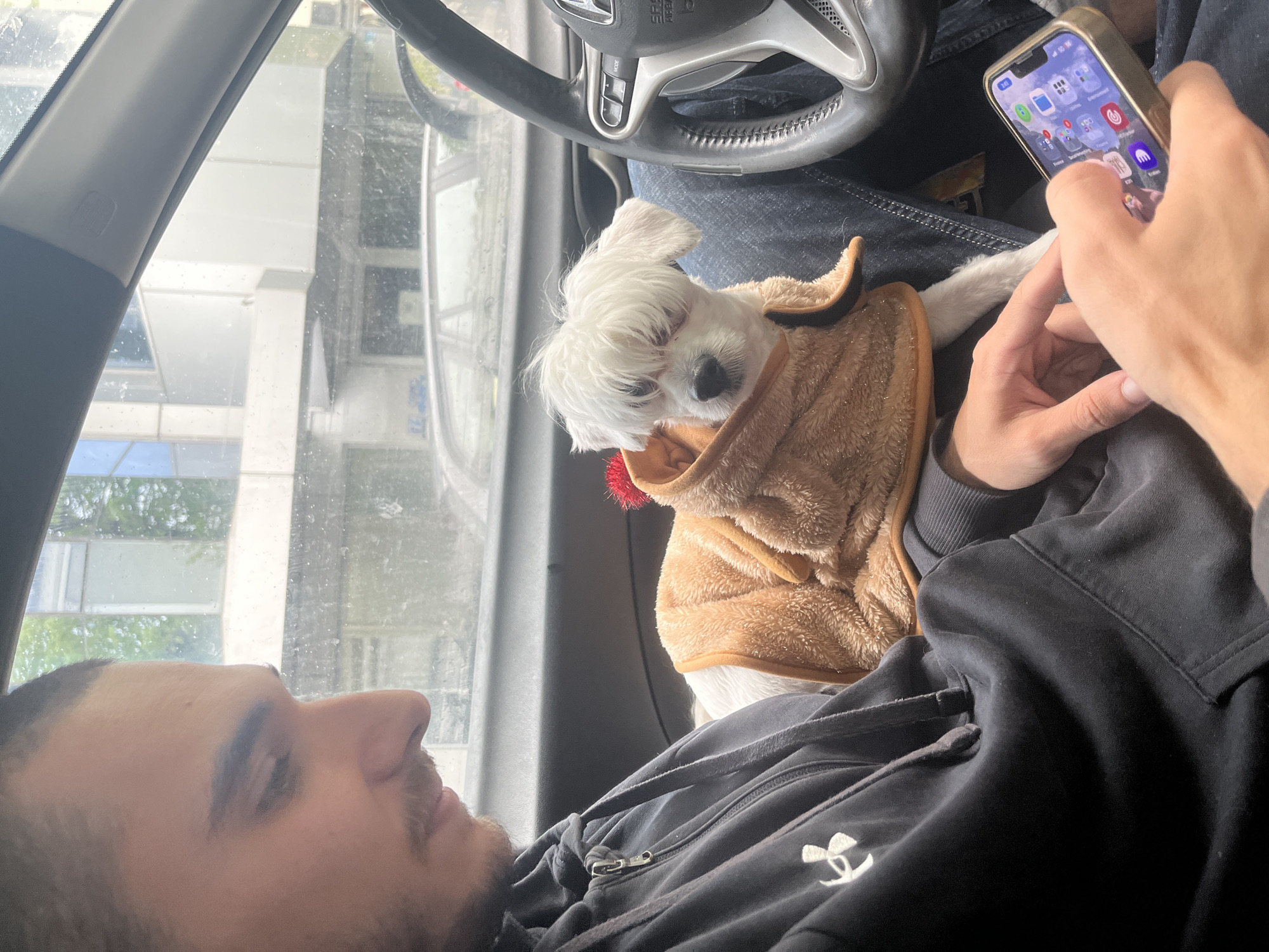 The image captures a relaxed scene inside a car, oriented as if the camera was rotated 90 degrees clockwise. It features a man, a small dog, and a person's hand using a smartphone, set within the car's interior.
The man, seen from the chest up in left profile, appears to be resting or looking out of the window. He has dark hair, a beard, and a mustache, and is wearing a black hoodie with a white Under Armour logo on the chest. A small, white, fluffy dog, possibly a Maltese, is nestled comfortably on what appears to be the man's lap or chest. The dog is wearing a cozy, brown, fuzzy hooded jacket, resembling a bear or reindeer costume with a small red pom-pom on top. Its eyes are partially closed, suggesting it is resting or dozing.
On the right side of the frame, a hand holds an iPhone, displaying its home screen with various app icons. Part of the car's steering wheel, featuring a "Honda" logo and "SRS AIRBAG" text, is visible behind the phone. More "SRS AIRBAG" text is also seen on the car's A-pillar. The view through the car's front window shows blurry reflections and outlines of buildings and trees, suggesting an urban setting. The window appears wet or dirty, obscuring the outside view. The overall atmosphere is calm and comfortable, indicating a leisurely moment, possibly while parked or stopped. The scene takes place in Varna, Bulgaria.