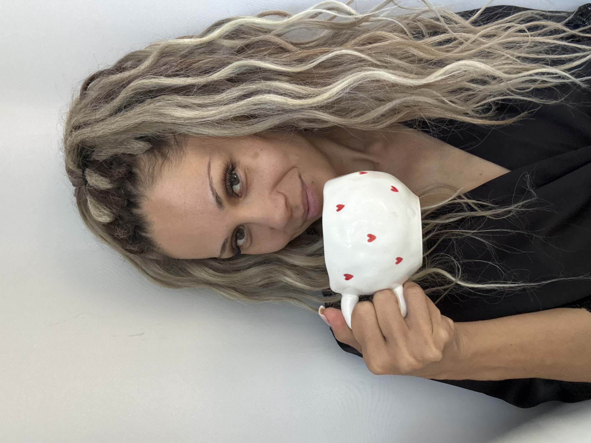 A woman is holding a white mug decorated with red hearts, brought to her lips in the middle of a selfie. Her light brown hair is styled into dreadlocks and cascades down her shoulders. She is wearing a black garment, possibly a robe or a dress with a deep neckline. The background is a plain, light-colored wall. No specific details about the city of Rusanovo, Russia, are visible in the image.