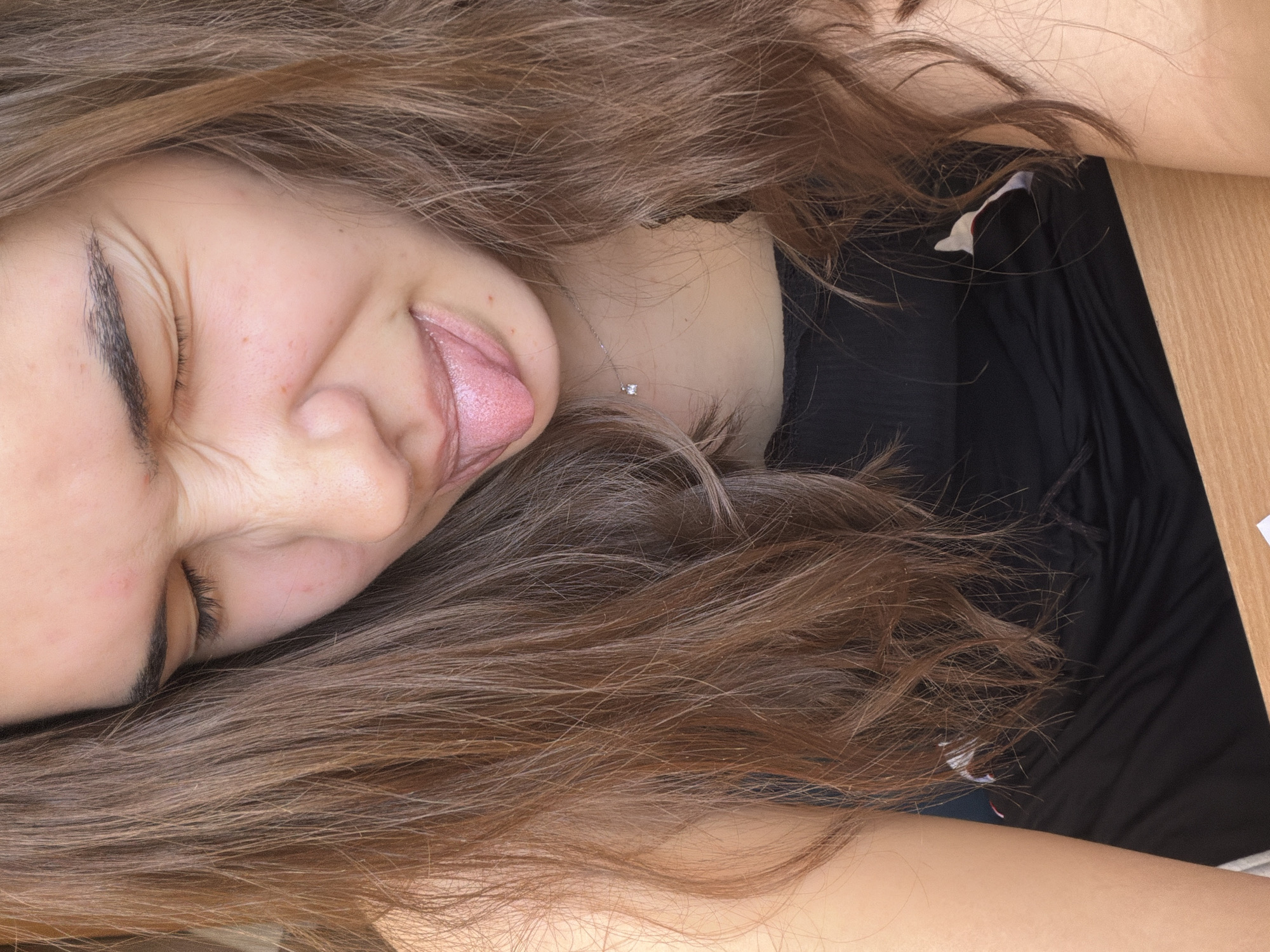 A young woman with light brown hair is lying on her side. Her eyes are closed and she is sticking out her tongue. She is wearing a black top and a delicate necklace with a single pendant. The background appears to be a wooden surface. The location is not discernible from the image.