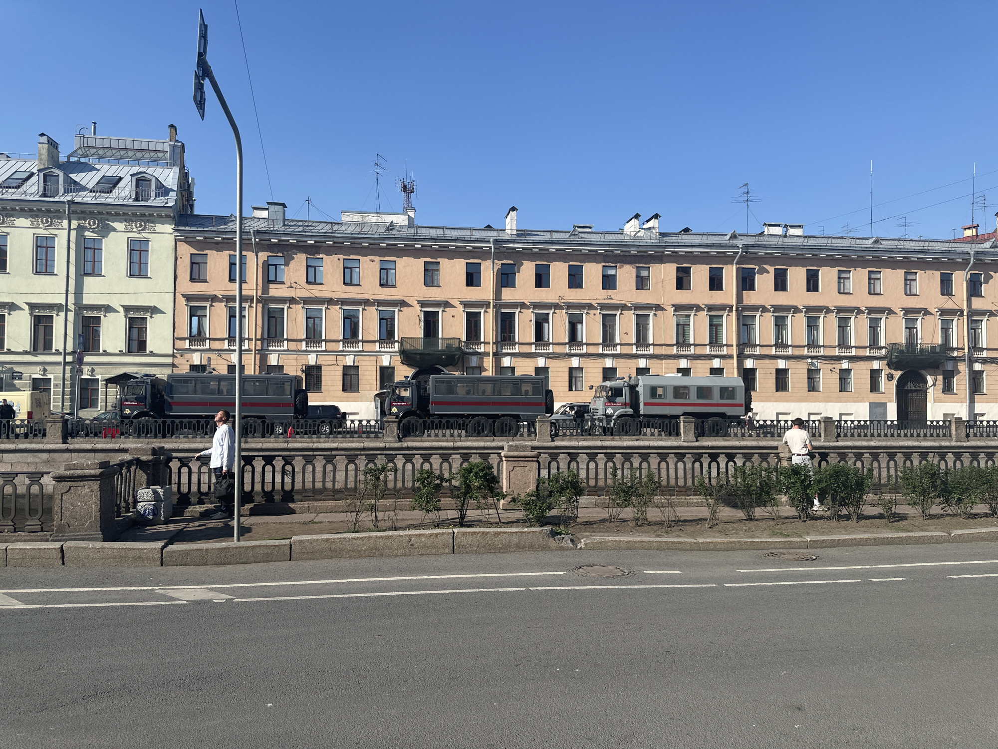 The image depicts a street scene in Saint Petersburg, Russia, characterized by architectural and infrastructural elements. A row of buildings stretches across the background, displaying diverse architectural styles. A pedestrian walkway, bordered by a stone balustrade and vegetation, runs parallel to the street. Several military trucks with passenger compartments are parked along the walkway behind the balustrade. Two individuals are present along the walkway, one dressed in white and another with a light-colored shirt, standing near the balustrade. The foreground is dominated by an asphalt road marked with white lines, under a clear, blue sky.