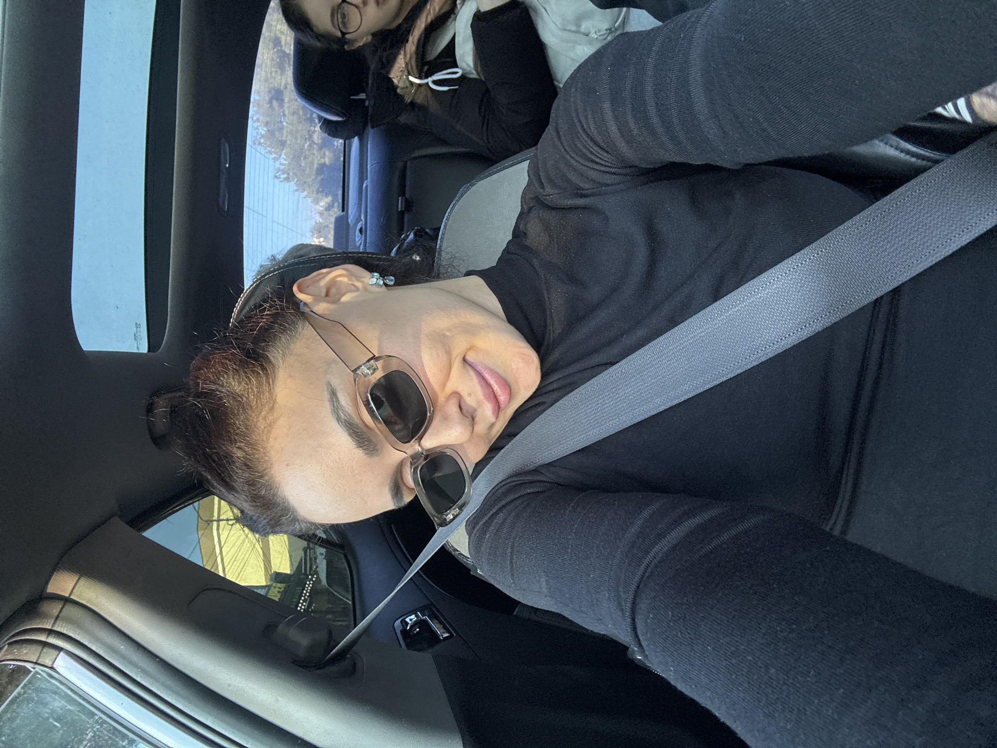 An adult female occupies the front passenger seat of a vehicle, angled towards the camera with a slight smile and tilted head. She wears a black long-sleeved turtleneck shirt, rectangular light-framed dark sunglasses, and light-colored teardrop earrings. A gray seatbelt is secured diagonally across her torso. Her dark hair is styled in a bun. A younger individual, wearing glasses and a dark winter coat with white cord details, is partially visible in the rear passenger seat directly behind her. The vehicle's interior is dark, featuring a visible chrome door handle on the left and a panoramic sunroof overhead, through which a clear blue sky is visible. Outside the left window, a bright yellow building and a dark sign partially displaying "UPER" are visible, accompanied by overhead power lines. Trees and a clear blue sky are visible through the rear window. This urban environment is consistent with the vicinity of an AgroHub supermarket in Tbilisi, Georgia.