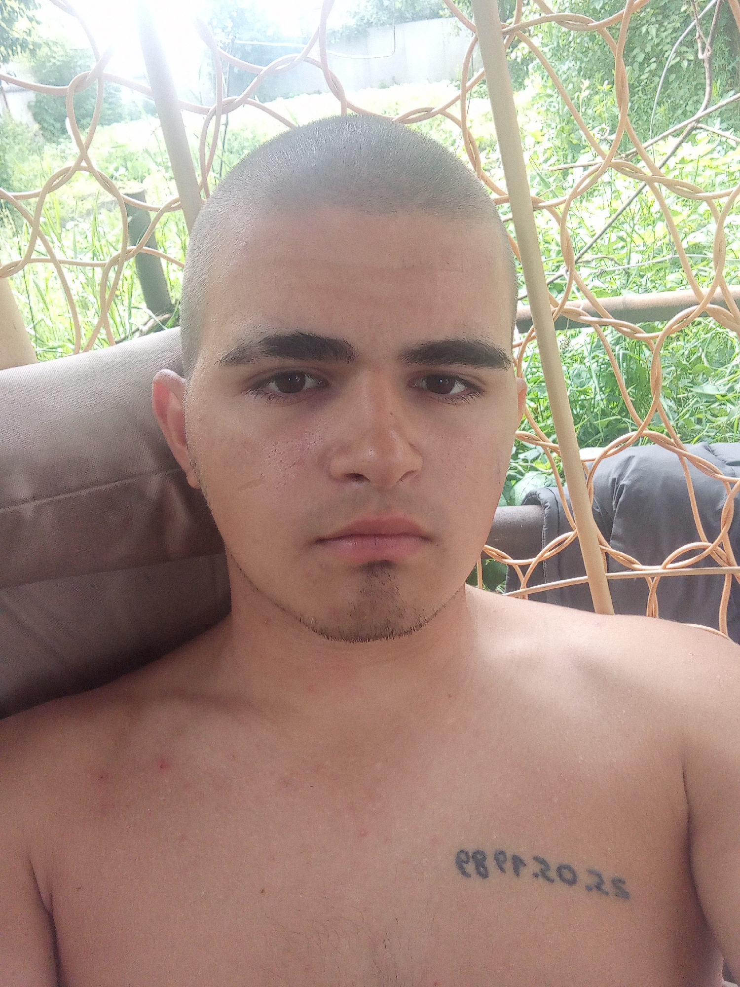 A young man with a shaved head and dark eyebrows is photographed from the chest up, seated outdoors. He is wearing a light-colored shirt, not visible in full, and has a tattoo on his chest. The background shows greenery, suggesting an outdoor setting. The image is likely taken in Kalach, Russia, based on the prompt.